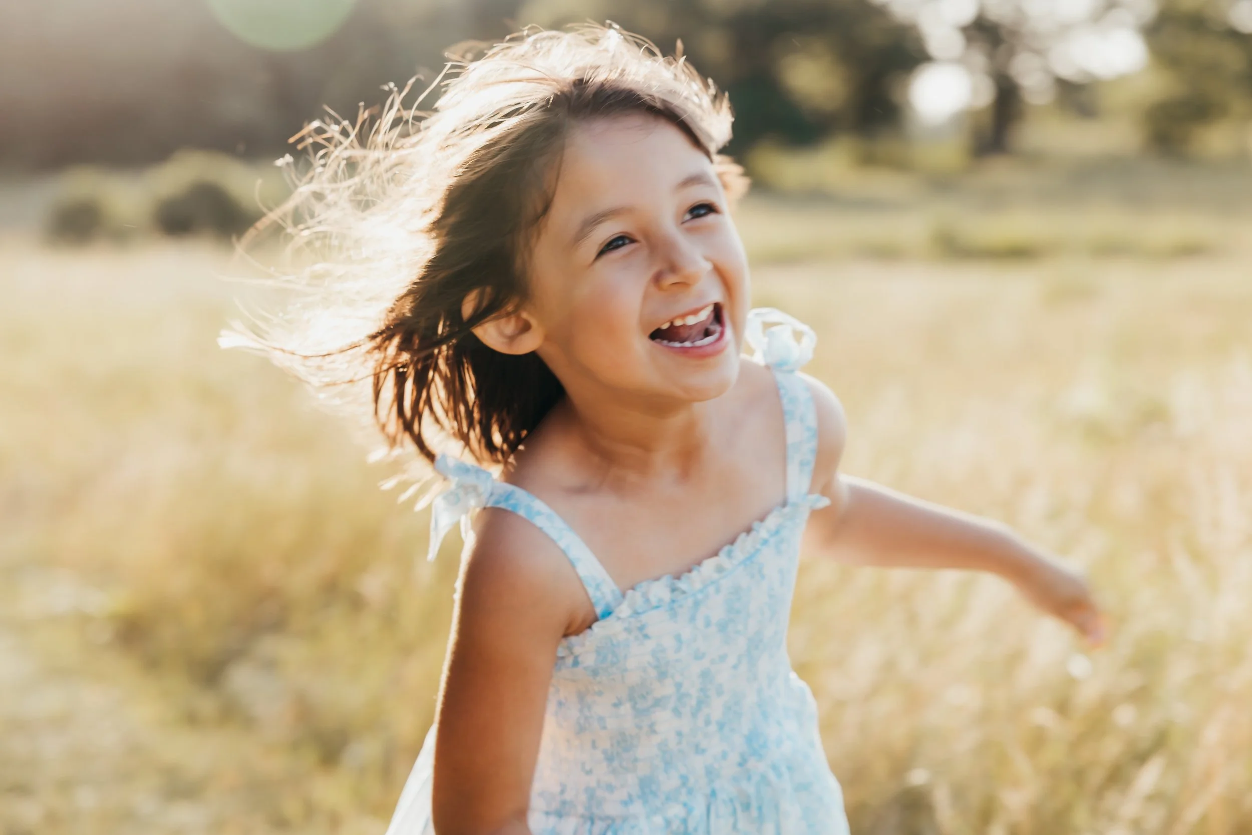 A young girl with dark hair wearing a light blue dress with white floral pattern, smiling happily outdoors in a sunny field with blurred trees in the background.