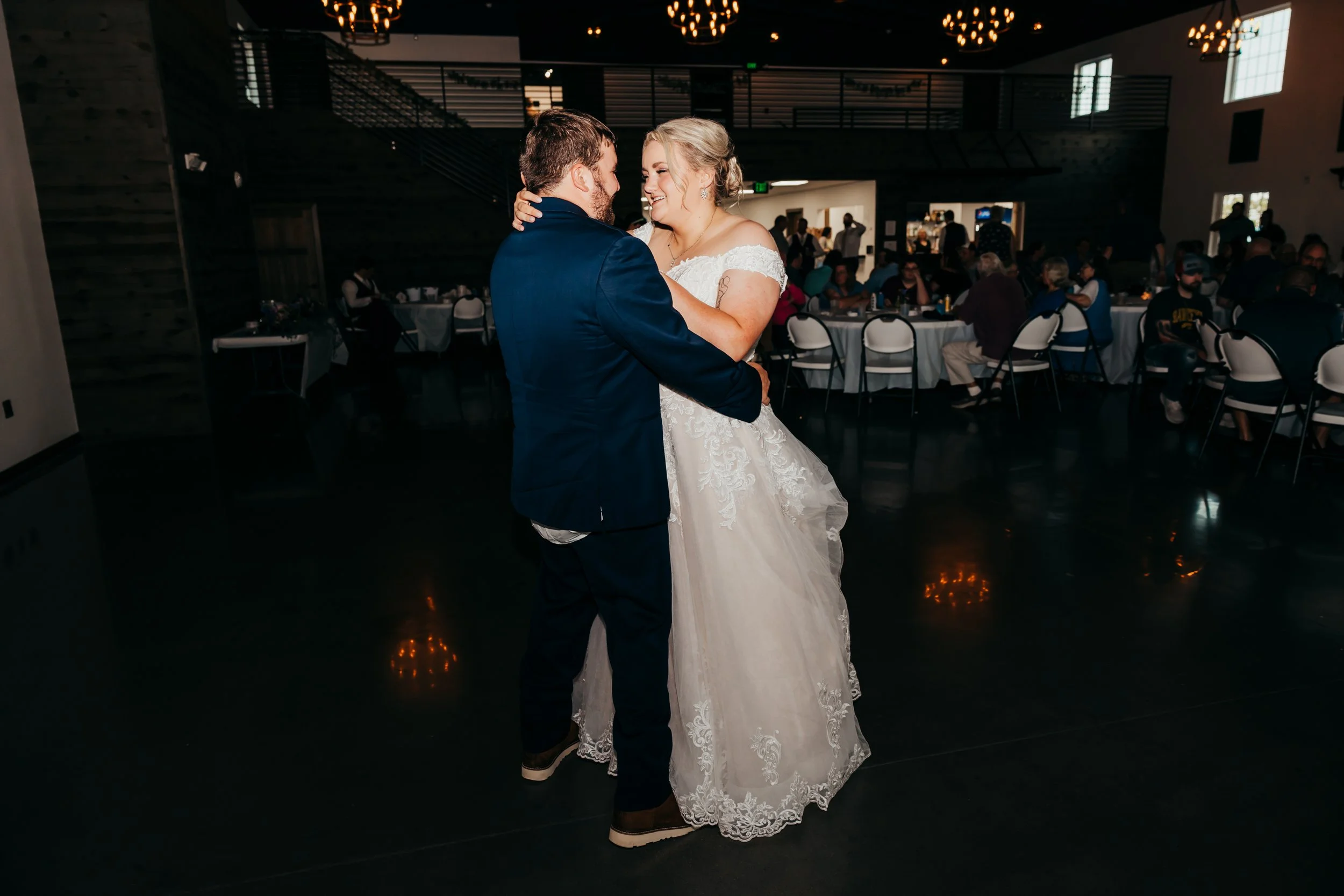 A bride and groom share their first dance at a wedding reception in a decorated indoor venue with guests seated at tables in the background.