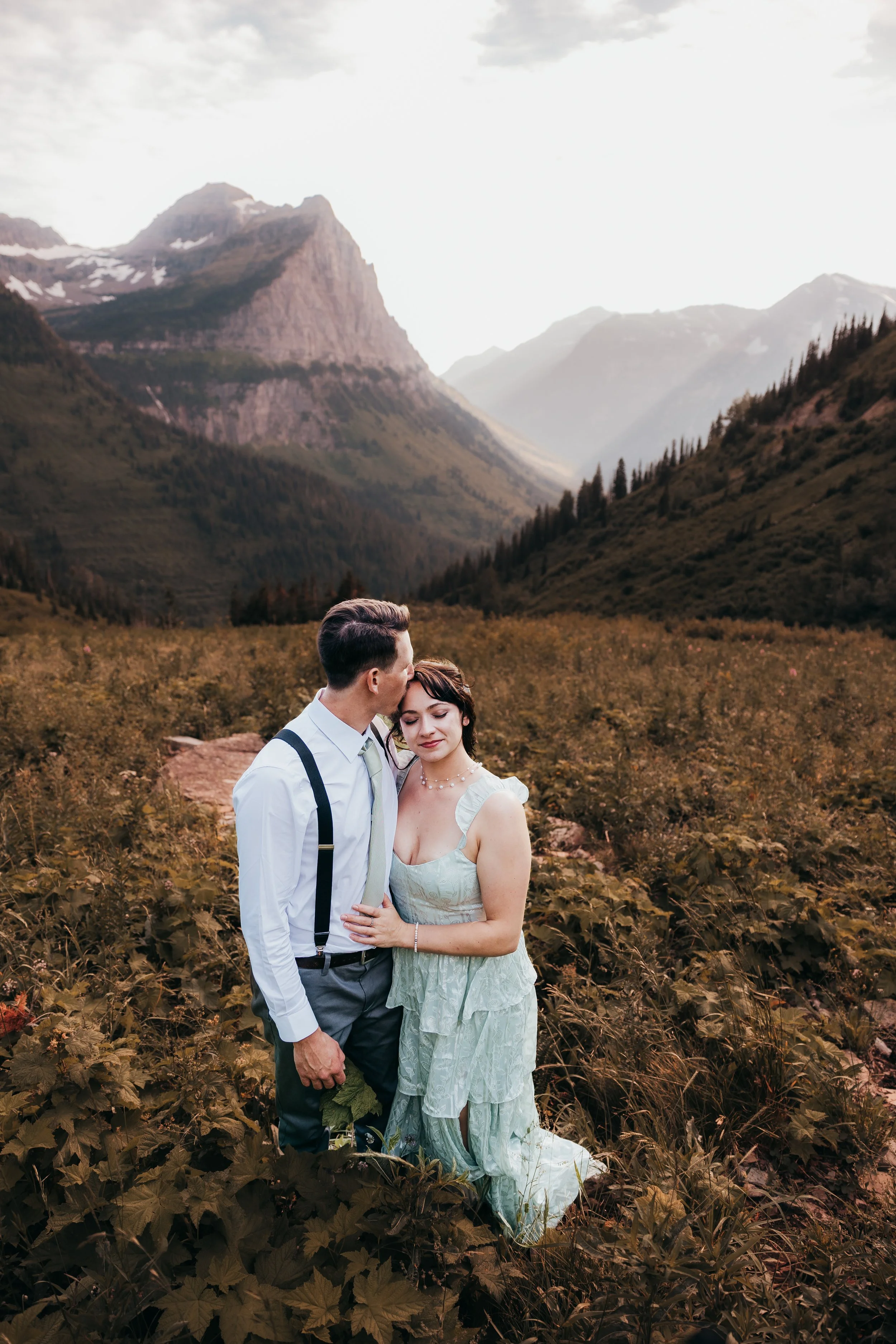 A couple standing close together outdoors in a mountainous area, with a man kissing a woman's forehead. The woman has dark hair and is wearing a light-colored dress, and the man has short hair and is dressed in a white shirt with suspenders. The back