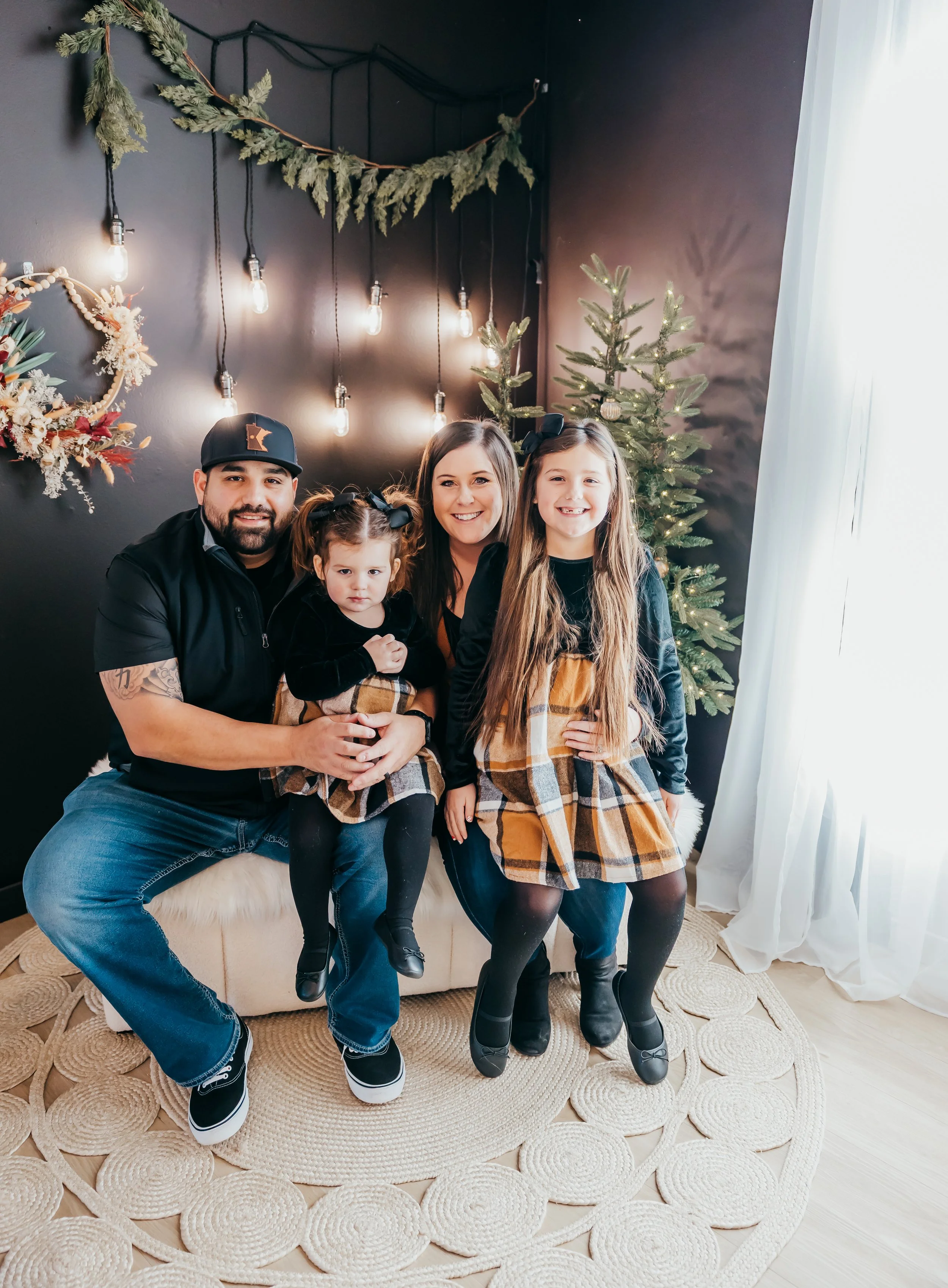 A family of four sitting on a beige ottoman in front of dark-colored wall decorated with hanging string lights, a wreath, and small Christmas trees. The family includes a man, woman, and two young girls, all smiling and dressed in black and plaid out