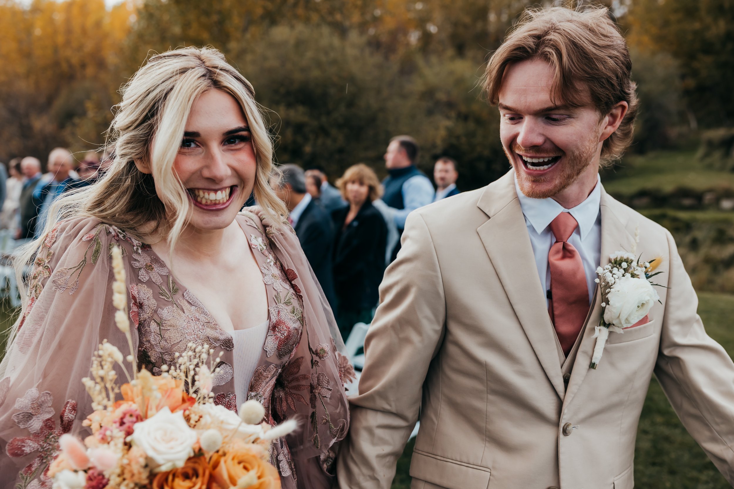 A smiling bride in a floral dress holding a bouquet, and a groom in a beige suit with a boutonniere, walking outdoors at a wedding celebration with guests and autumn trees in the background.