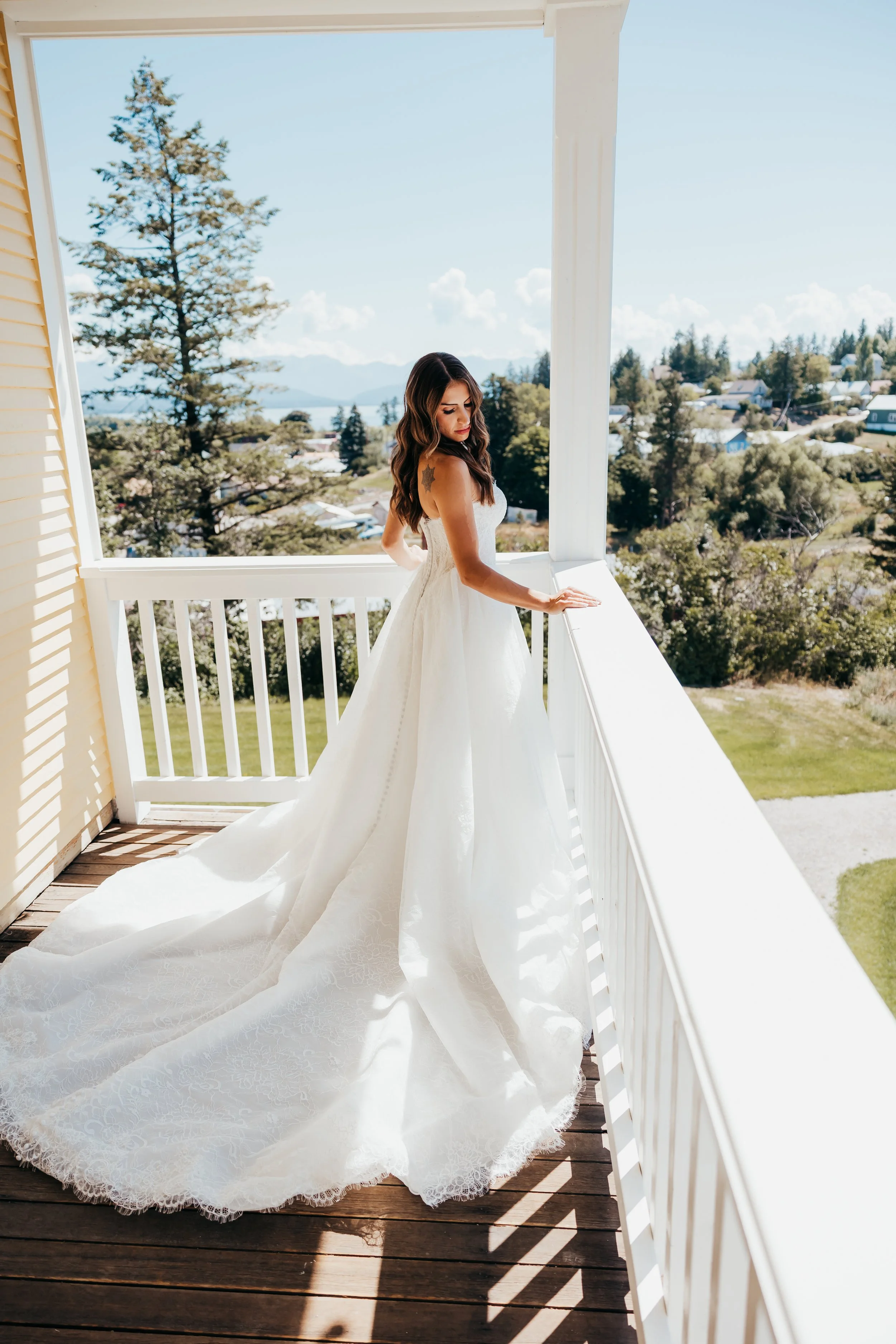 A woman in a white wedding dress standing on a balcony overlooking a scenic landscape with trees, houses, and mountains in the background.
