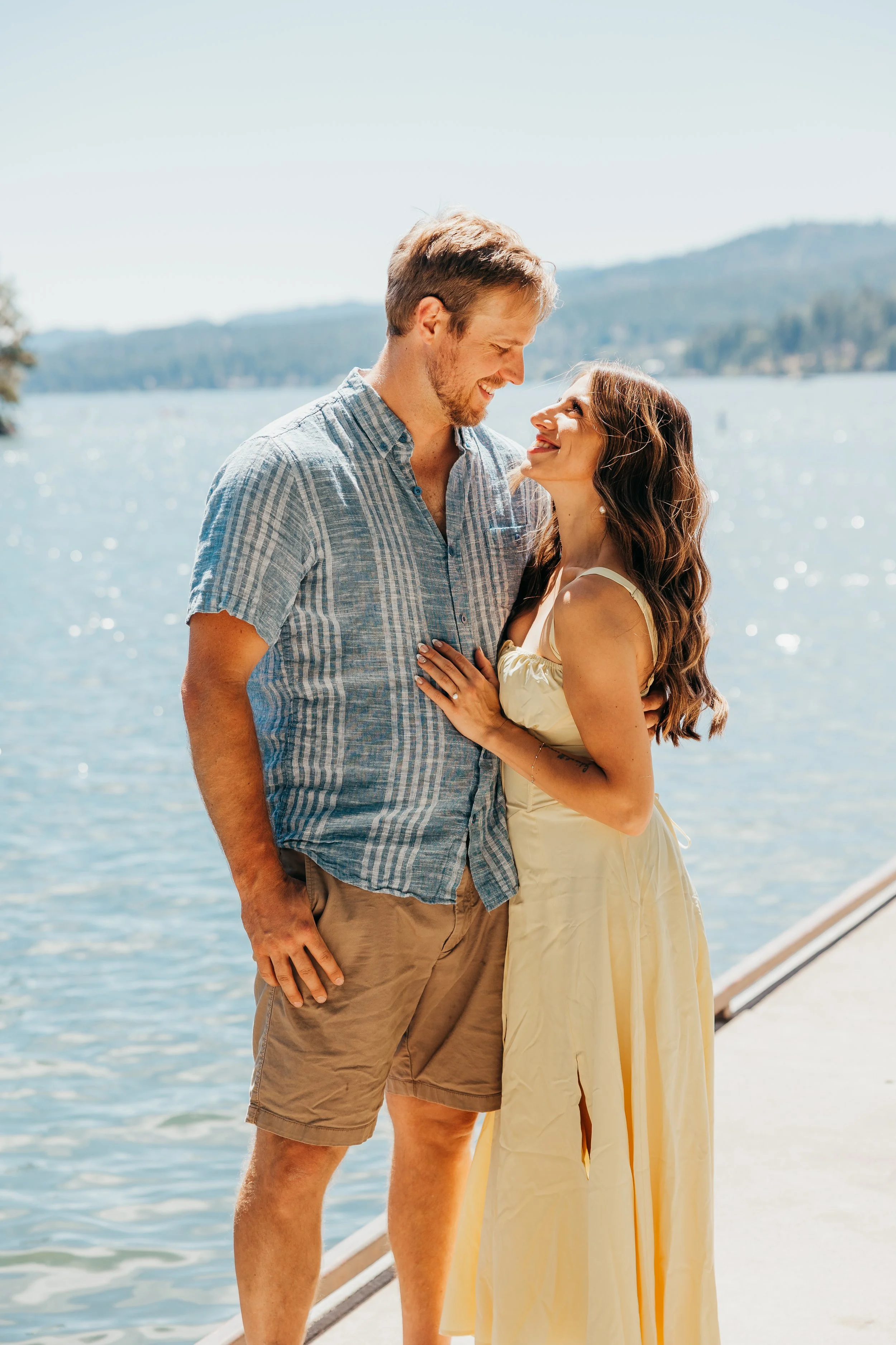 A couple smiling and looking at each other by the water, with a lake and mountains in the background on a sunny day.
