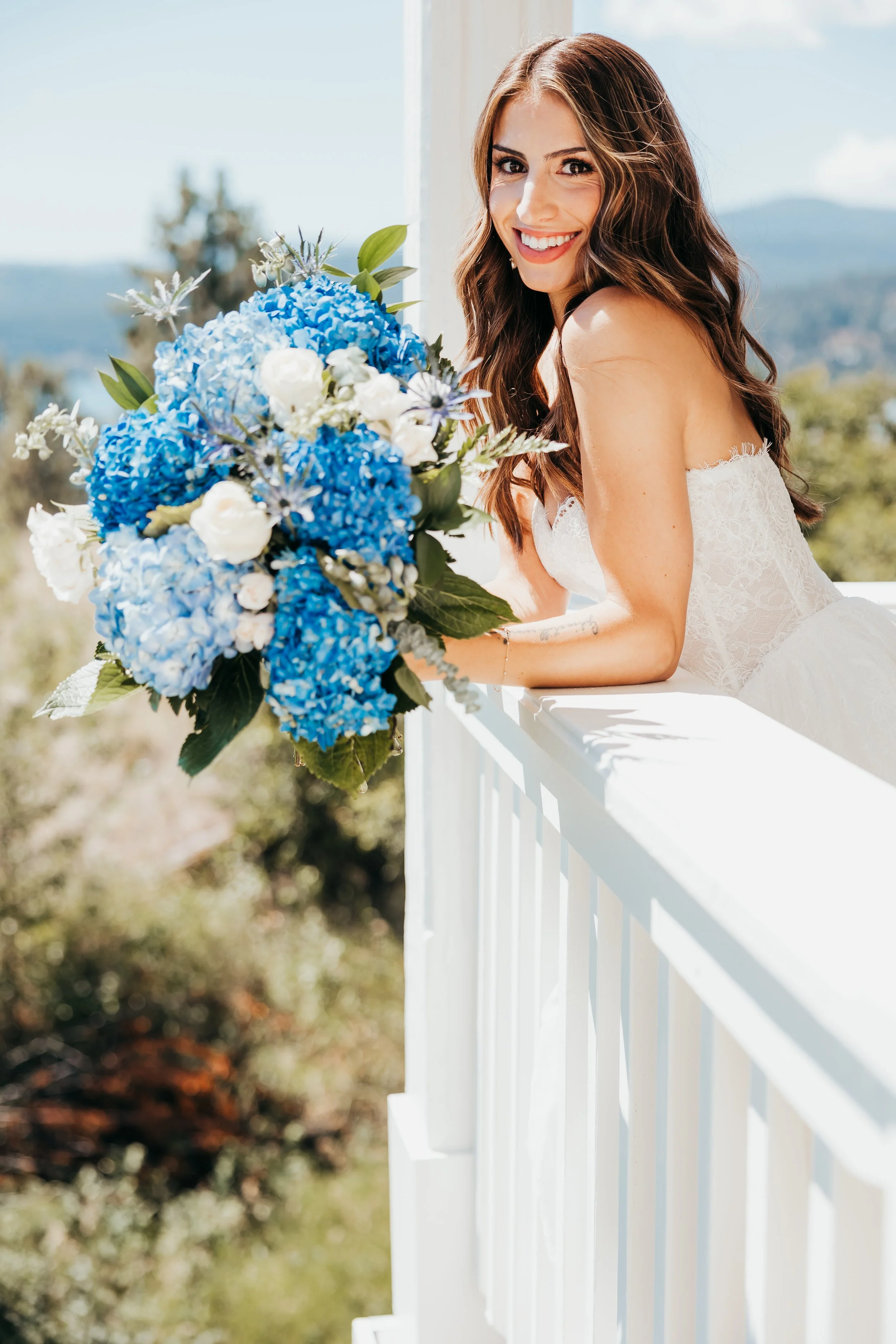 A smiling bride with long wavy brown hair in a strapless white wedding dress, leaning on a white railing, with a large blue and white floral bouquet, outdoors on a sunny day.