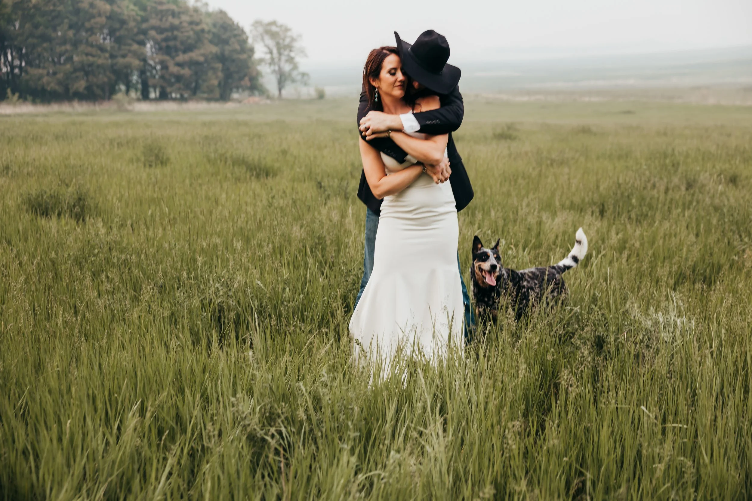 A couple embracing in a grassy field, with a dog standing nearby and trees and open land in the background.