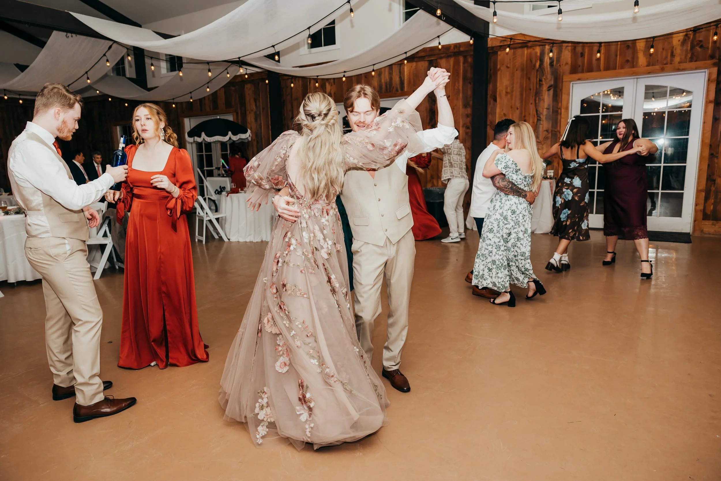 People dancing at a wedding reception in a rustic indoor venue with string lights and draped fabric decorations.