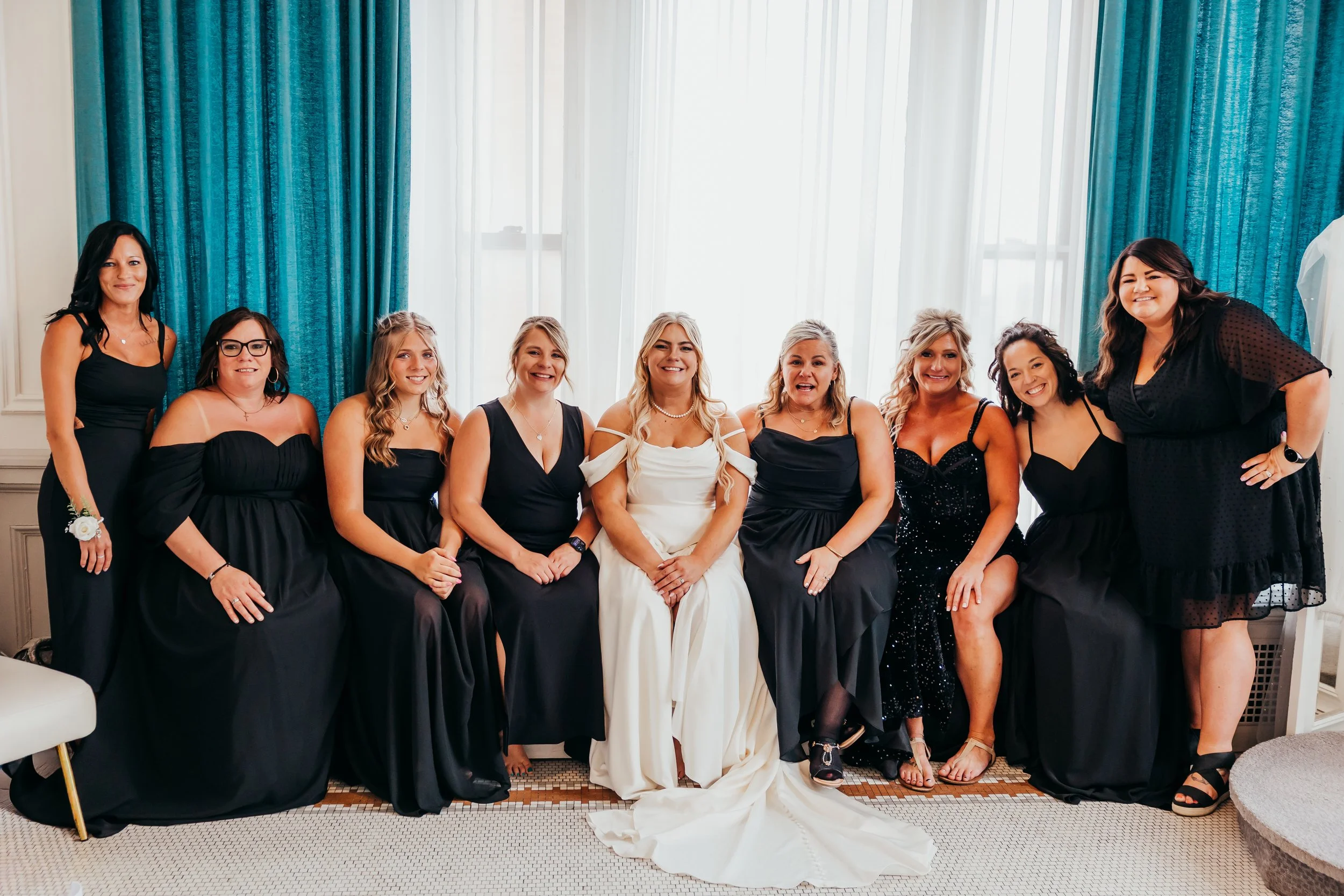 Group of women, with one bride in white wedding dress, sitting and standing in front of blue curtains in a bright room, smiling for a photo.