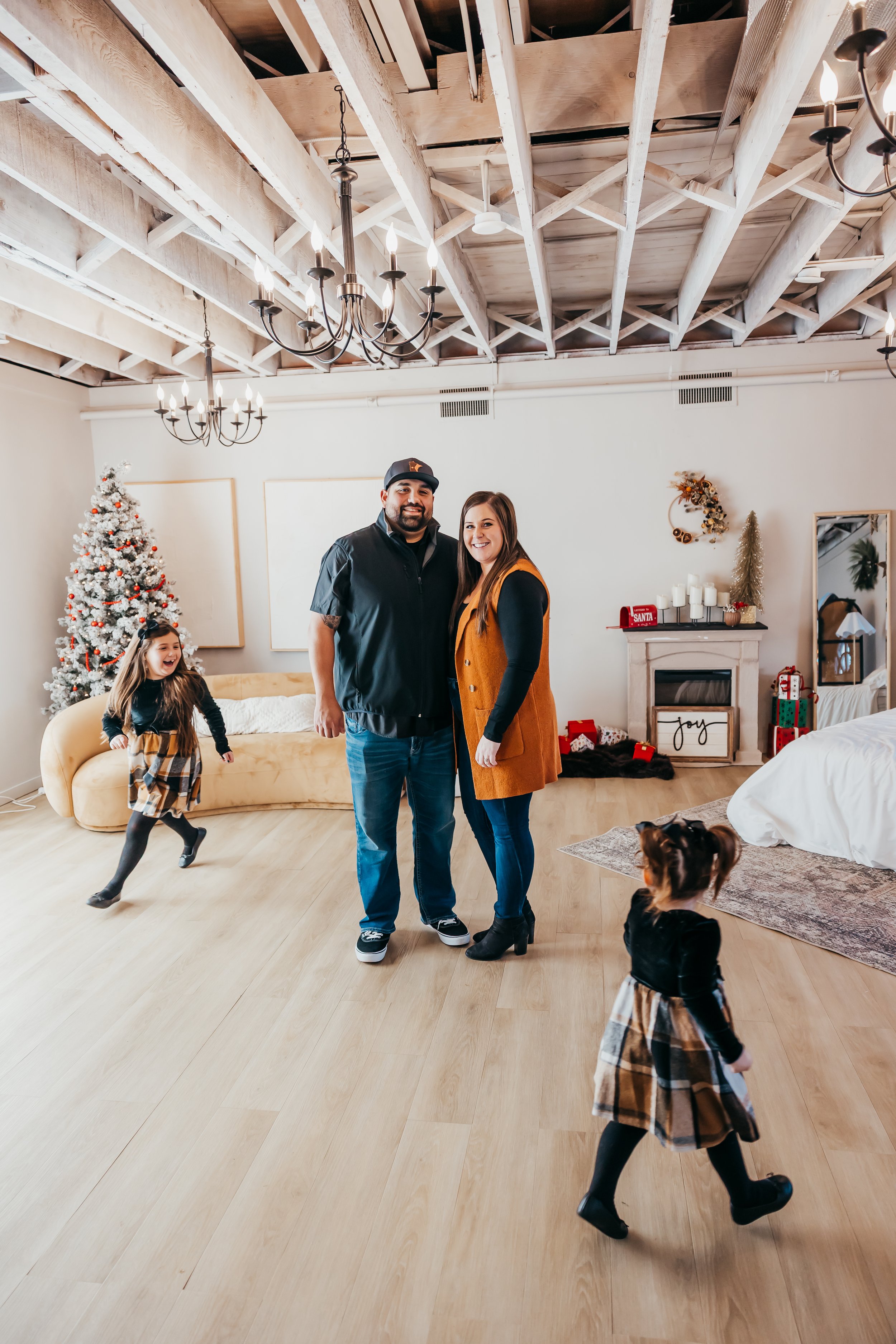 A family celebrating Christmas indoors, with a decorated Christmas tree, two adults, and two children in festive dresses, in a warmly decorated living room.