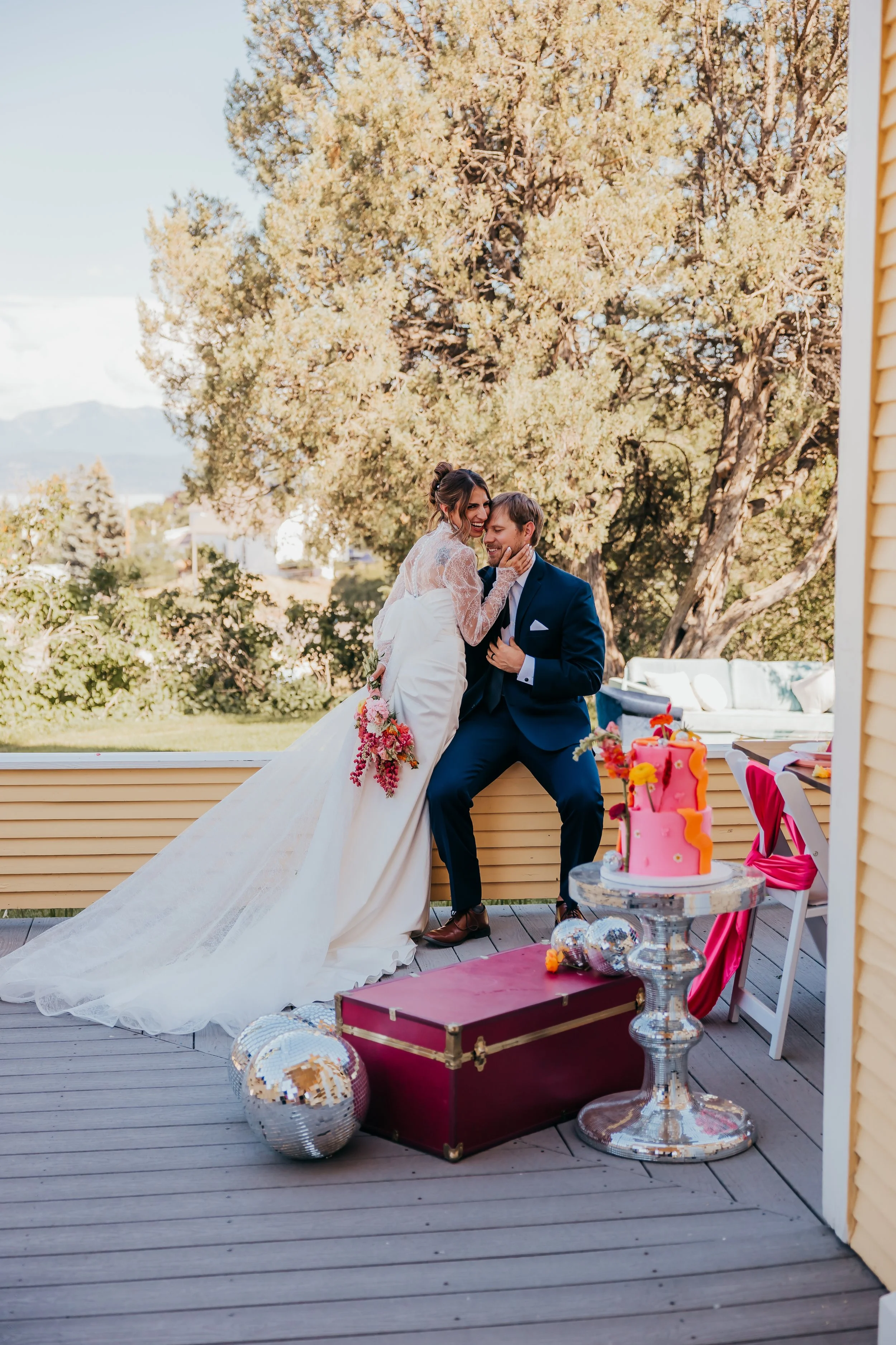 Bride and groom celebrating at their wedding outdoors, with a large tree and scenic background. The bride is holding a bouquet, and the groom is sitting on a ledge, both smiling and leaning towards each other.