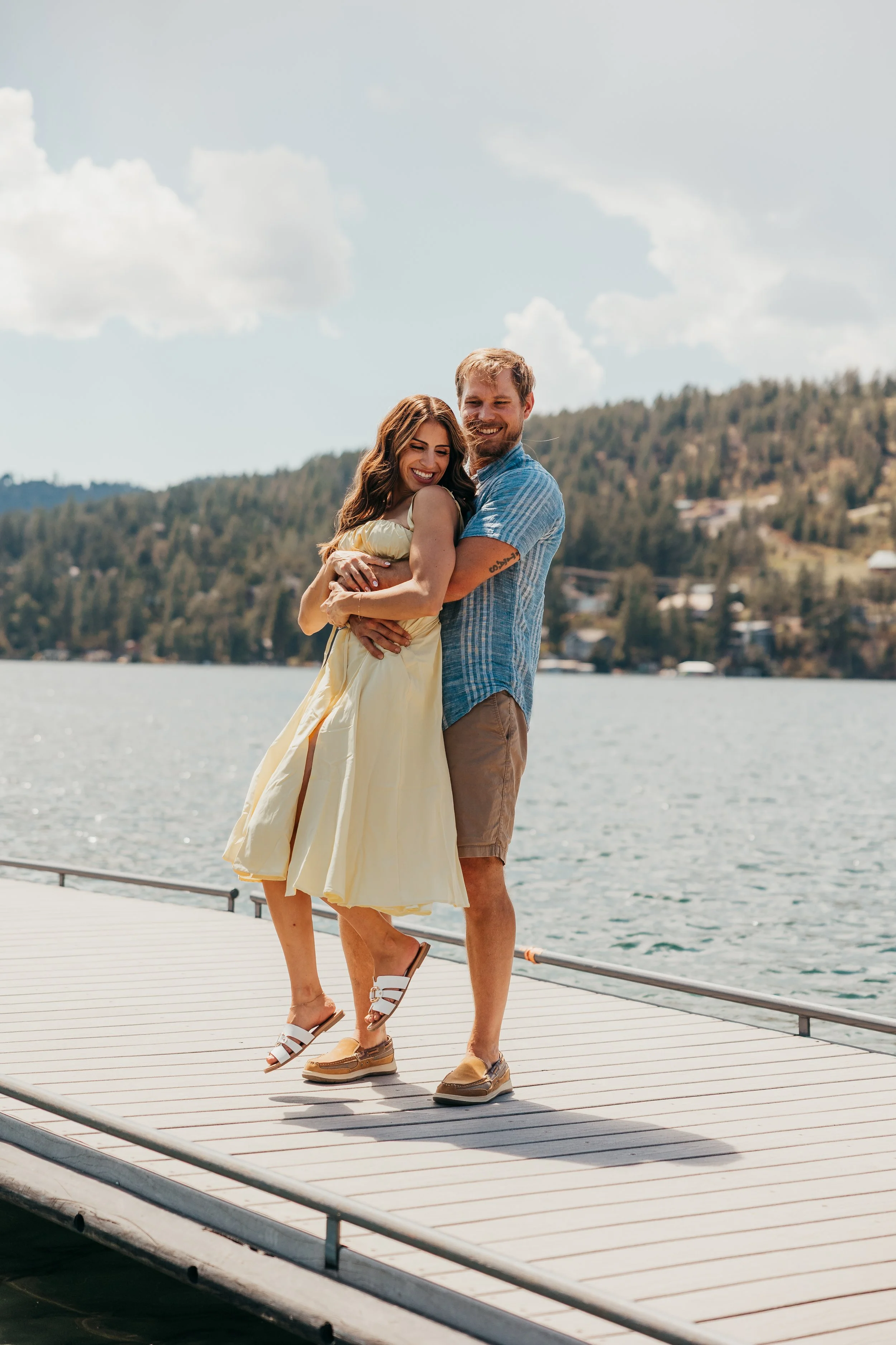 A young couple hugging and smiling on a dock by a lake with trees and hills in the background on a sunny day.