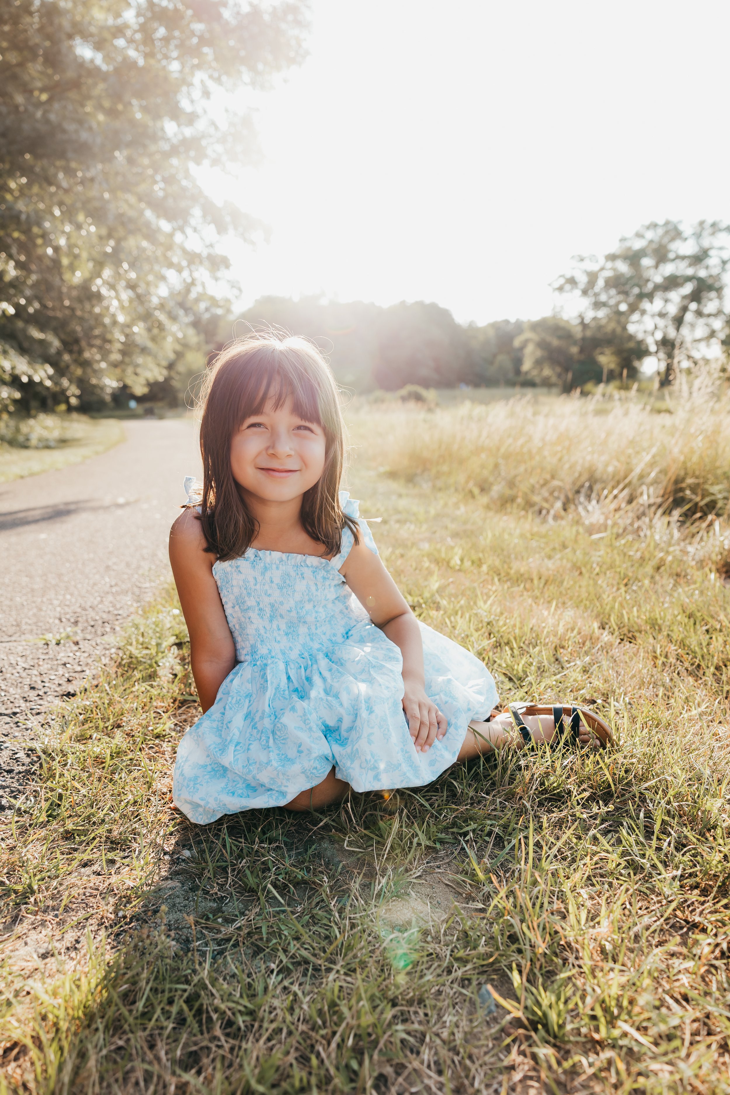 A young girl with brown hair and a blue and white dress sitting outdoors on grass, smiling at the camera, with sunlight and trees in the background.