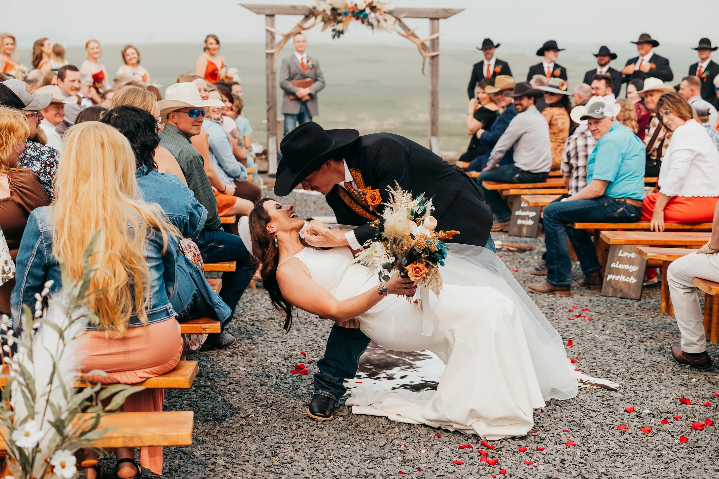 A bride and groom share a moment during their outdoor wedding ceremony, with guests seated on wooden benches and a decorated arch in the background.