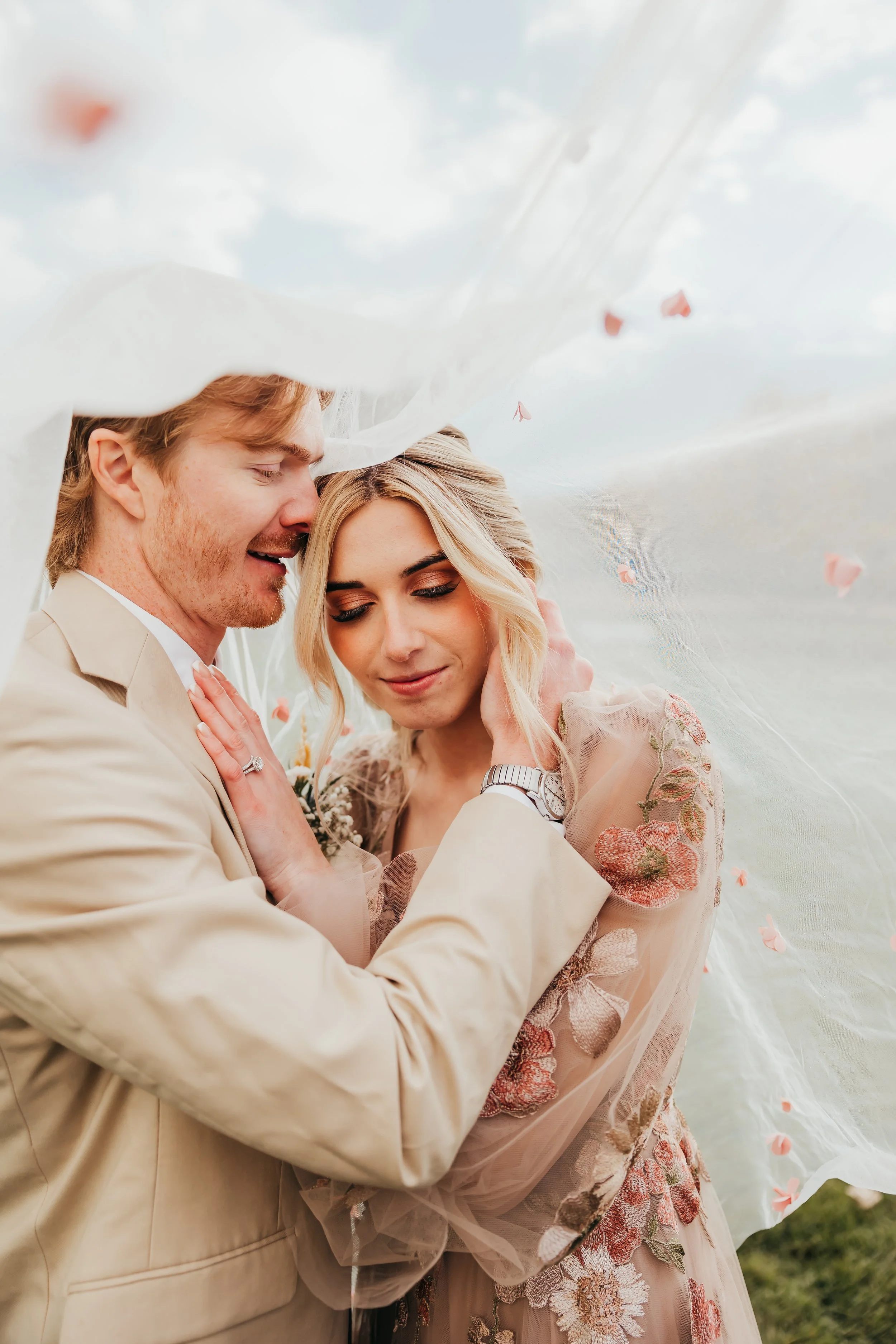 A romantic moment between a bride and groom surrounded by fabric with pink flower petals, outdoors under a cloudy sky.