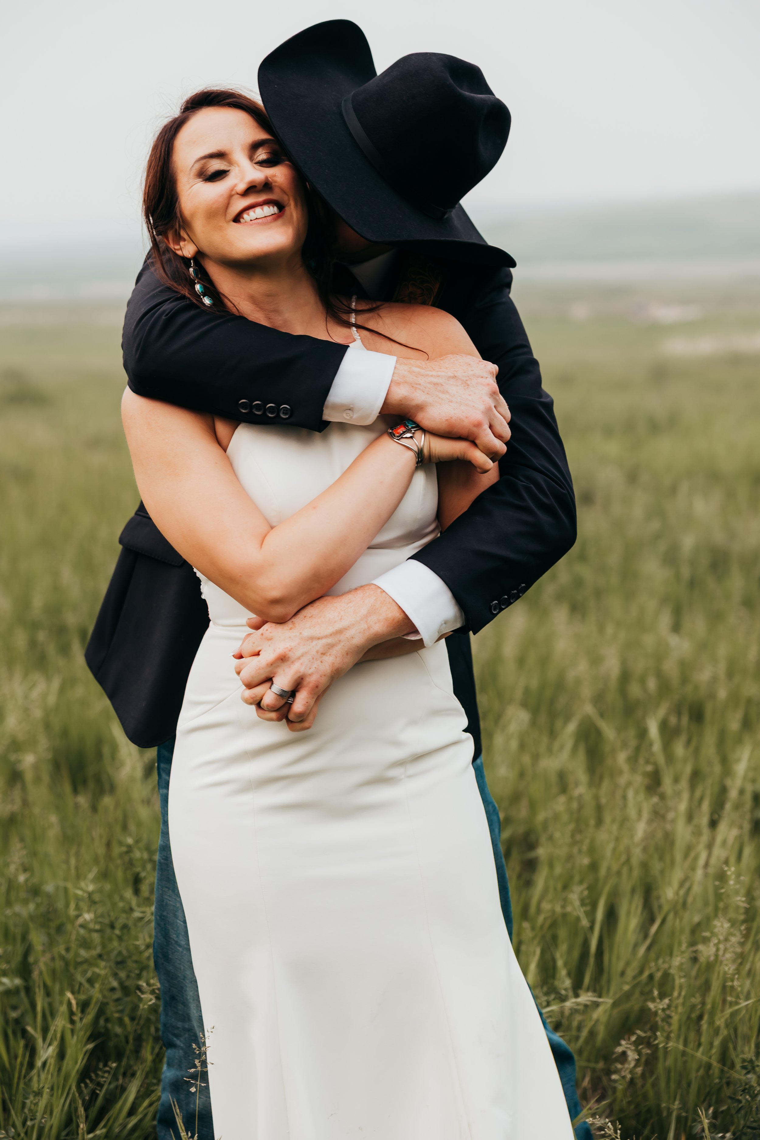 A woman in a white dress and a man in a black hat and suit embrace and hug in a grassy field.