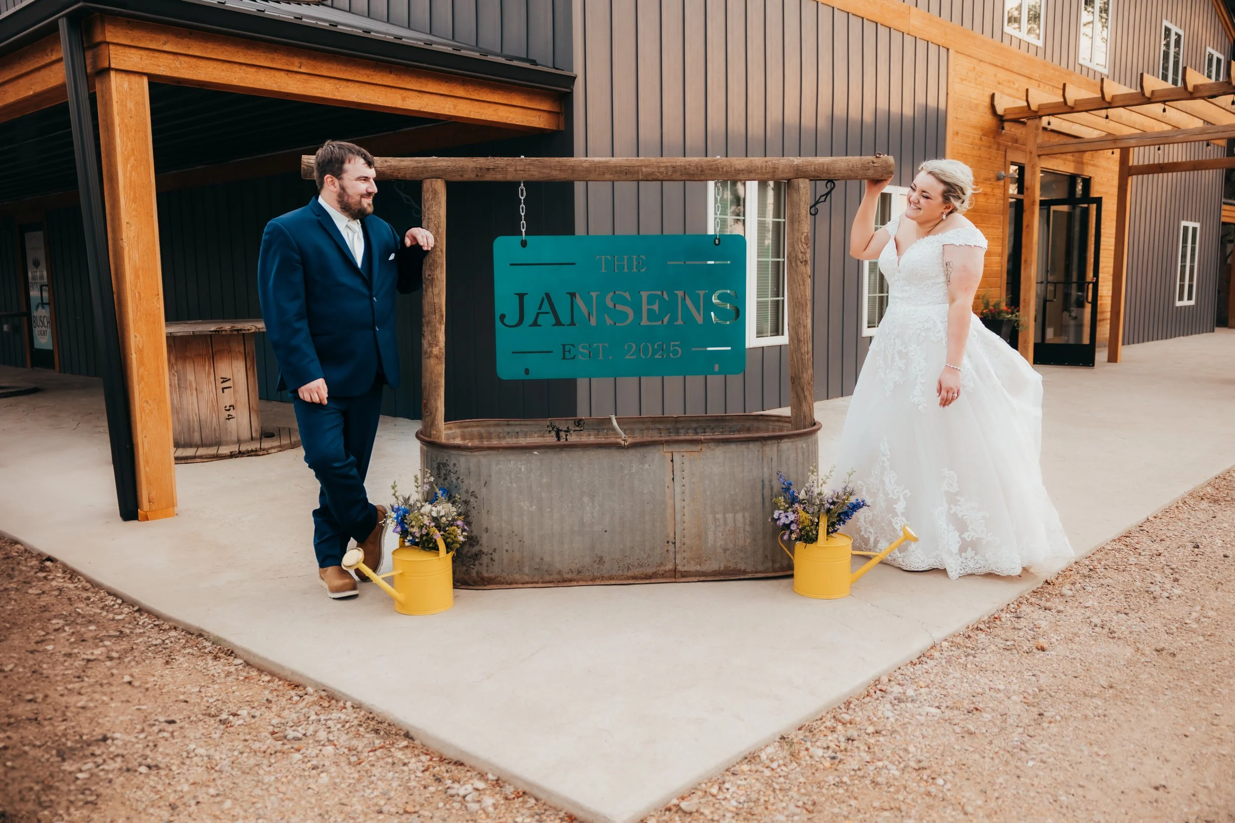 A bride and groom in wedding attire standing outside near a sign that reads "The Jansens Est. 2025," with colorful flower arrangements in yellow watering cans at the base.
