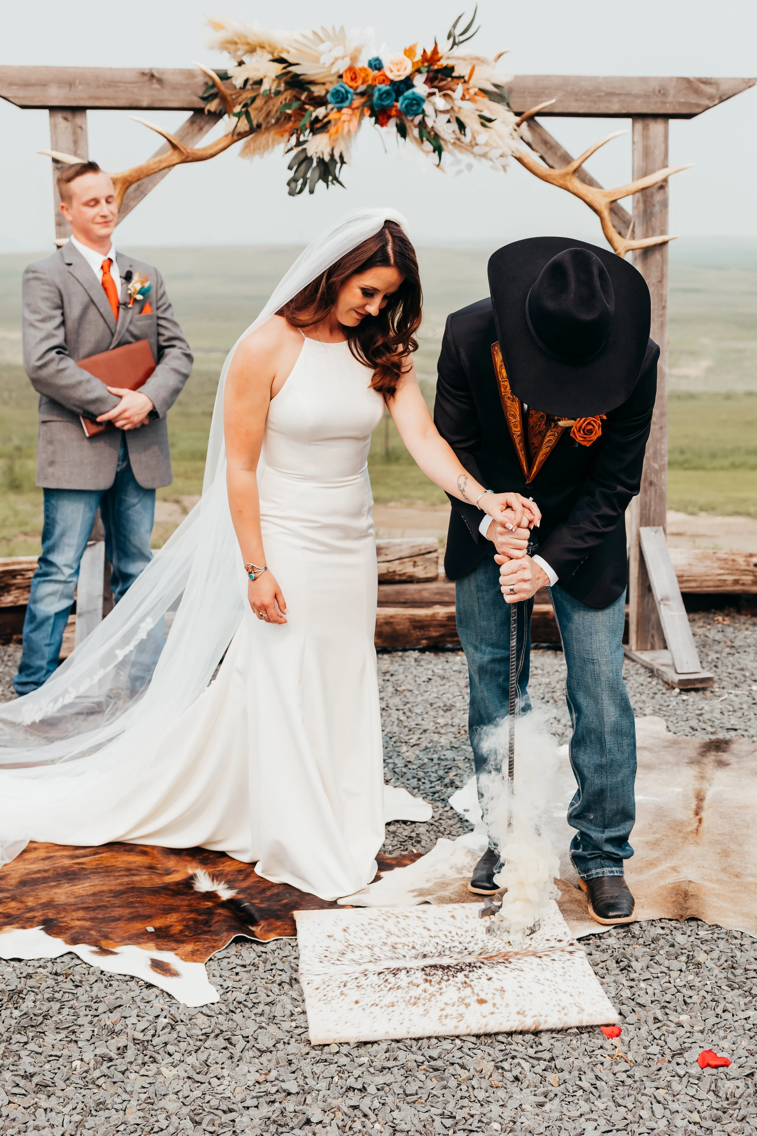 A couple at a rustic outdoor wedding ceremony, with the bride in a white gown and veil, the groom in a dark jacket with a hat, and an officiant standing behind them, lighting a fire on a small rug with animal hides on a gravel ground, under a wooden 