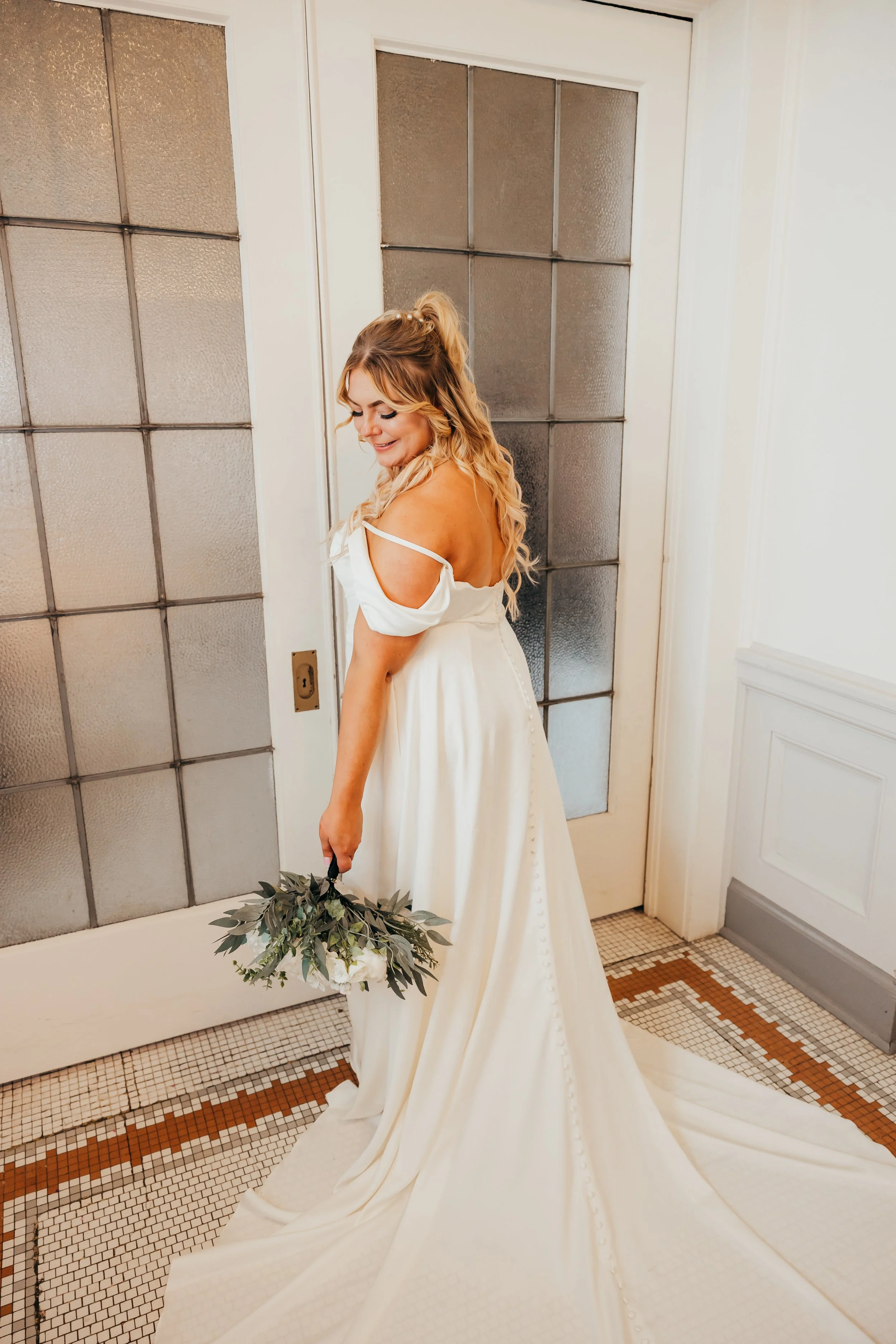 A bride in a white wedding gown holding a bouquet, standing indoors near a metal-framed glass door, smiling and looking down.