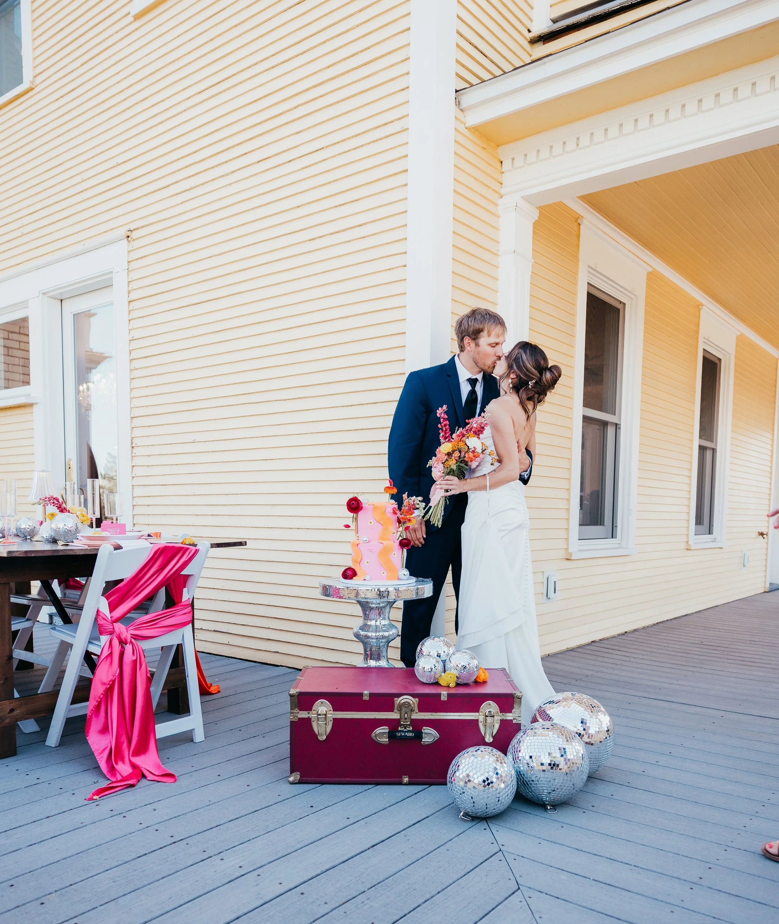 Bride and groom sharing a kiss at their outdoor wedding reception. The bride holds a colorful bouquet, and a wedding cake with pink and yellow ice cream decorations rests on a small table. Disco balls are scattered on the ground, with the reception a