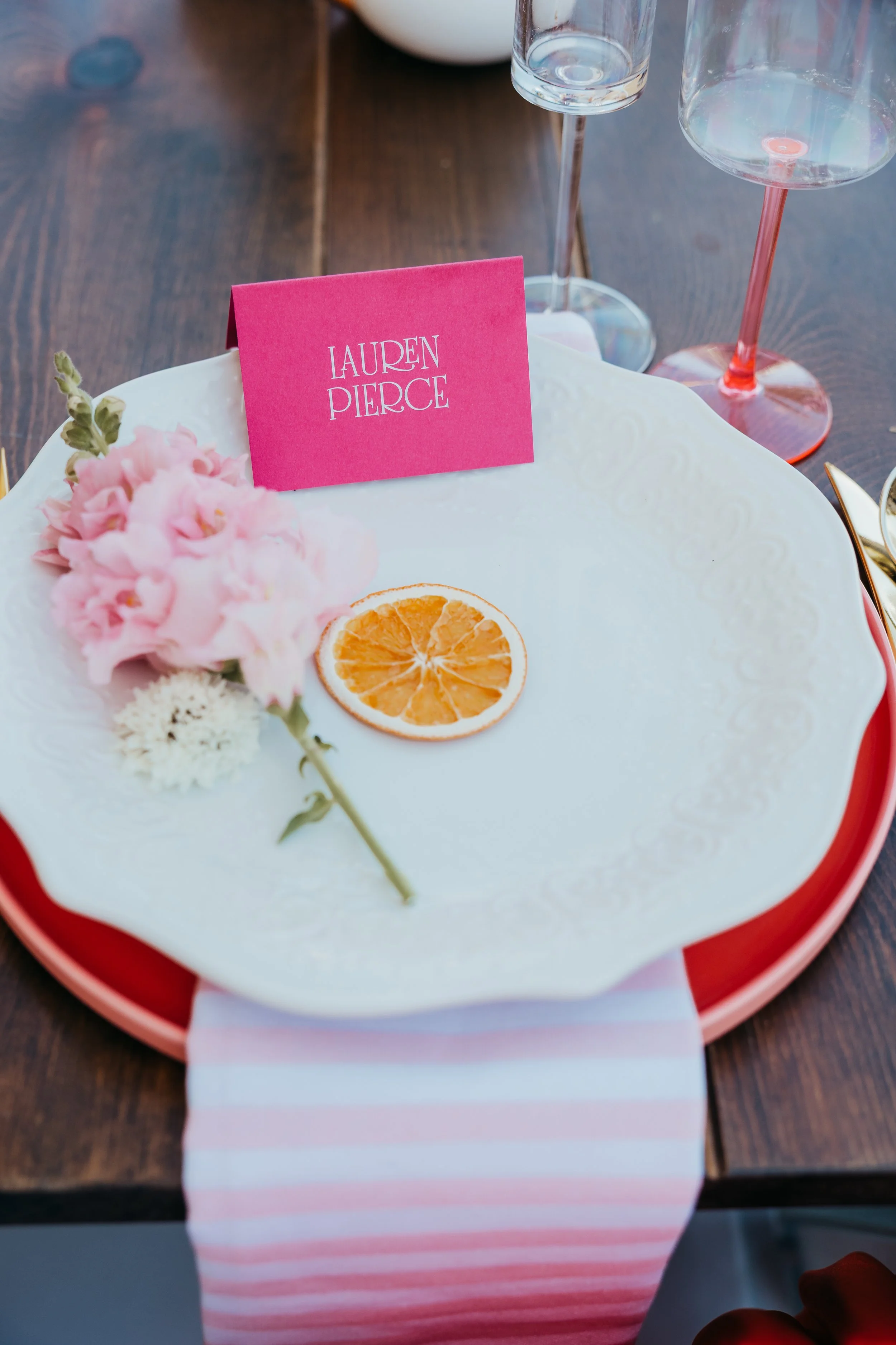 A white decorative plate with a pink place card labeled "LAUREN PIERCE," a slice of dried orange, pink flowers, and a white flower, set on a wooden table with a pink and white striped napkin underneath. Two glasses, one clear and one with a pink stem