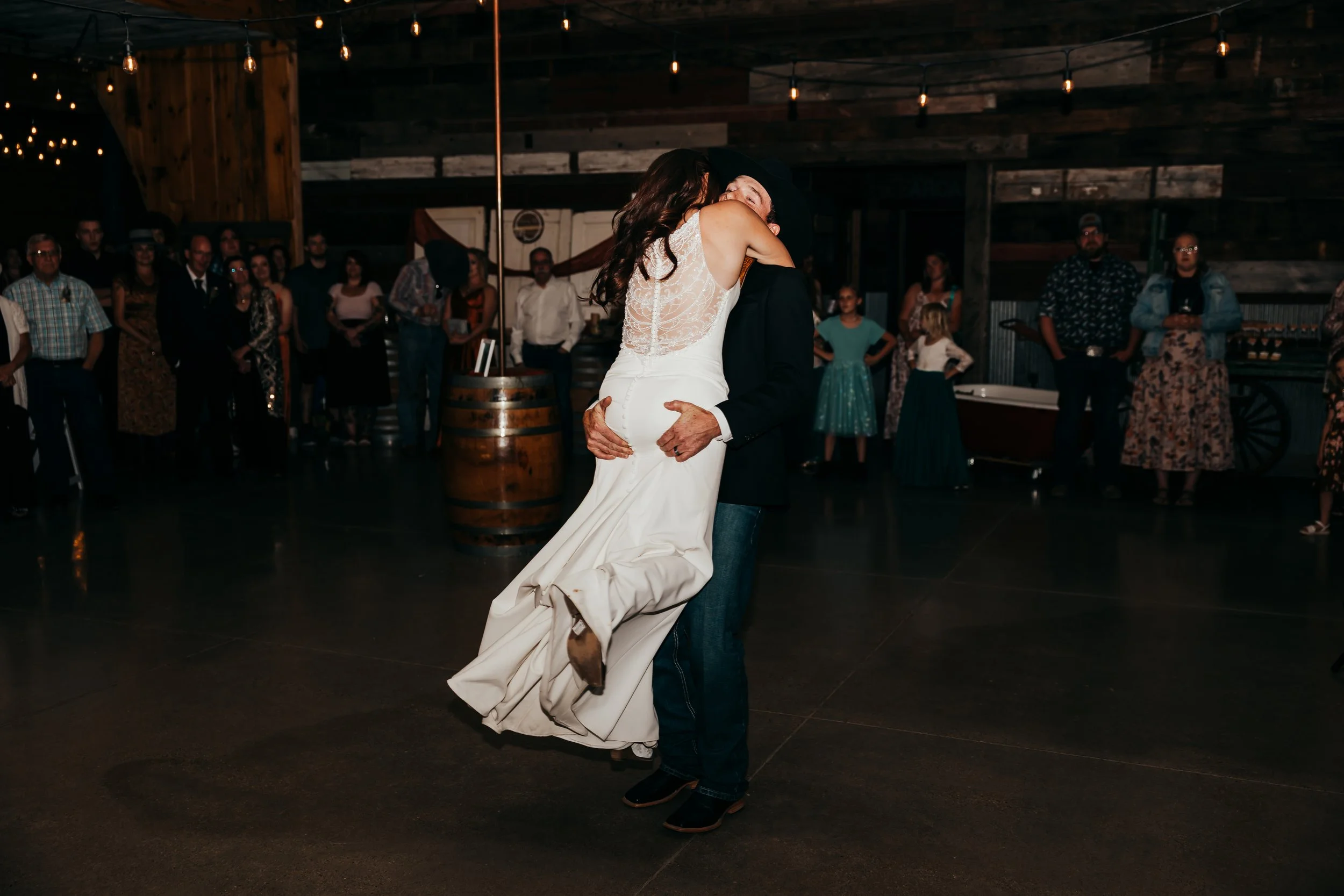 Couple sharing a dance at a wedding reception with guests watching in a rustic barn setting.