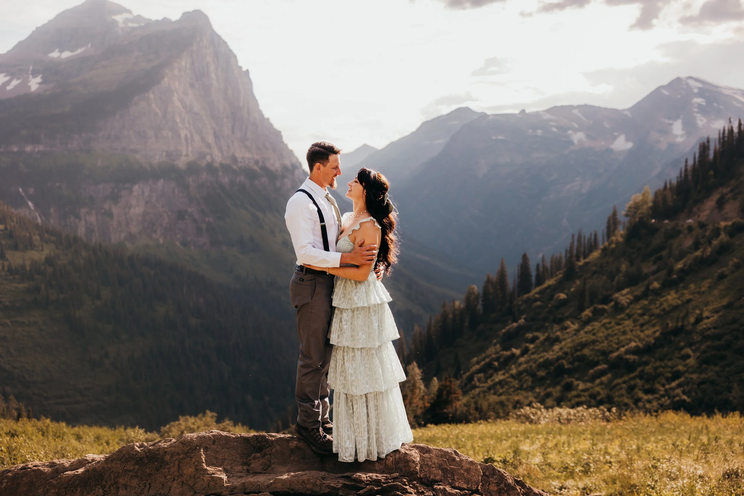 A couple in wedding attire standing on a rock in a mountainous landscape, embracing and looking at each other, with mountains and trees in the background.