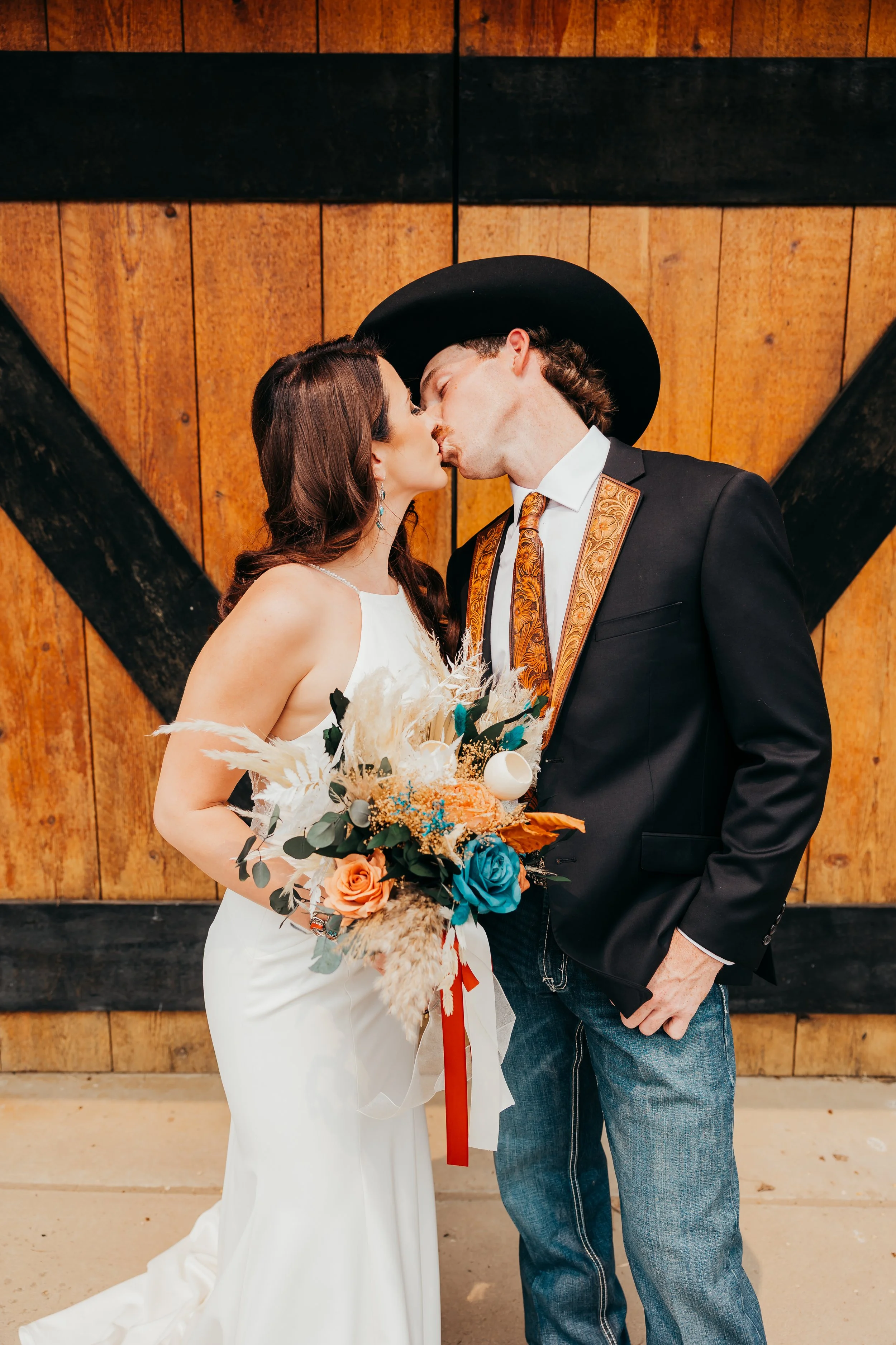 A bride and groom sharing a kiss indoors; the bride wears a white dress and holds a bouquet, and the groom is in a black jacket, a printed tie, and a large black hat, with a wooden wall background.