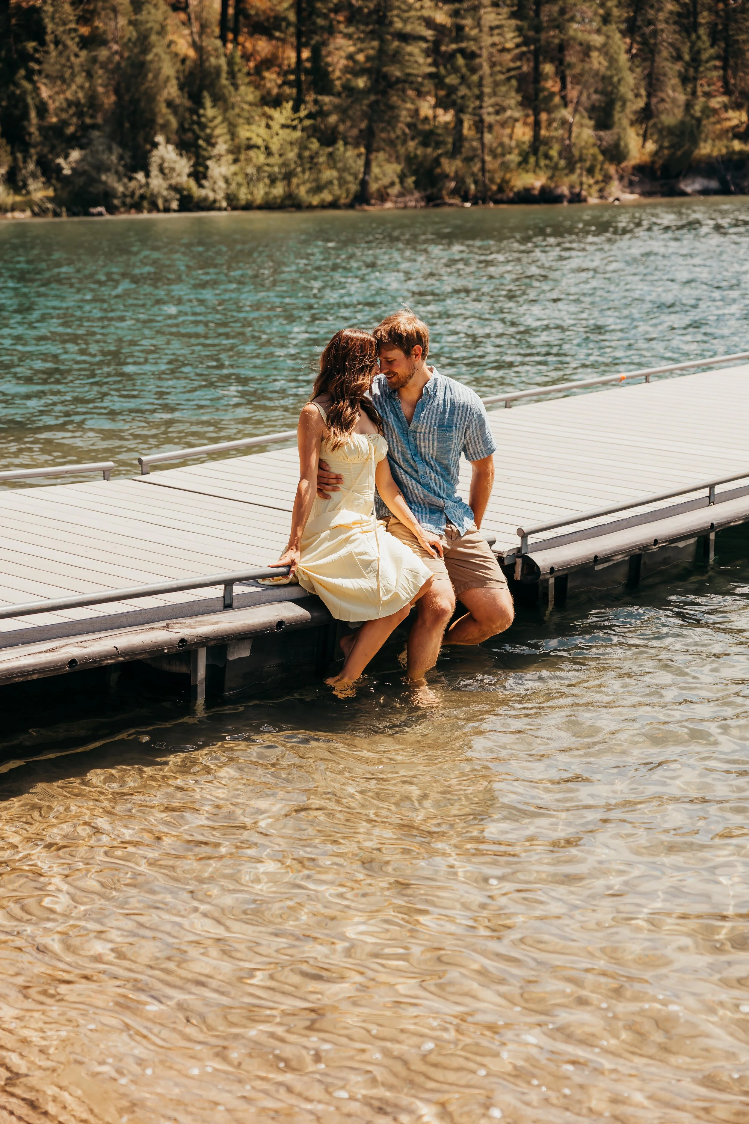 A couple sitting on a dock with their legs in the water, exchanging a kiss near a lake with a forested shoreline in the background.