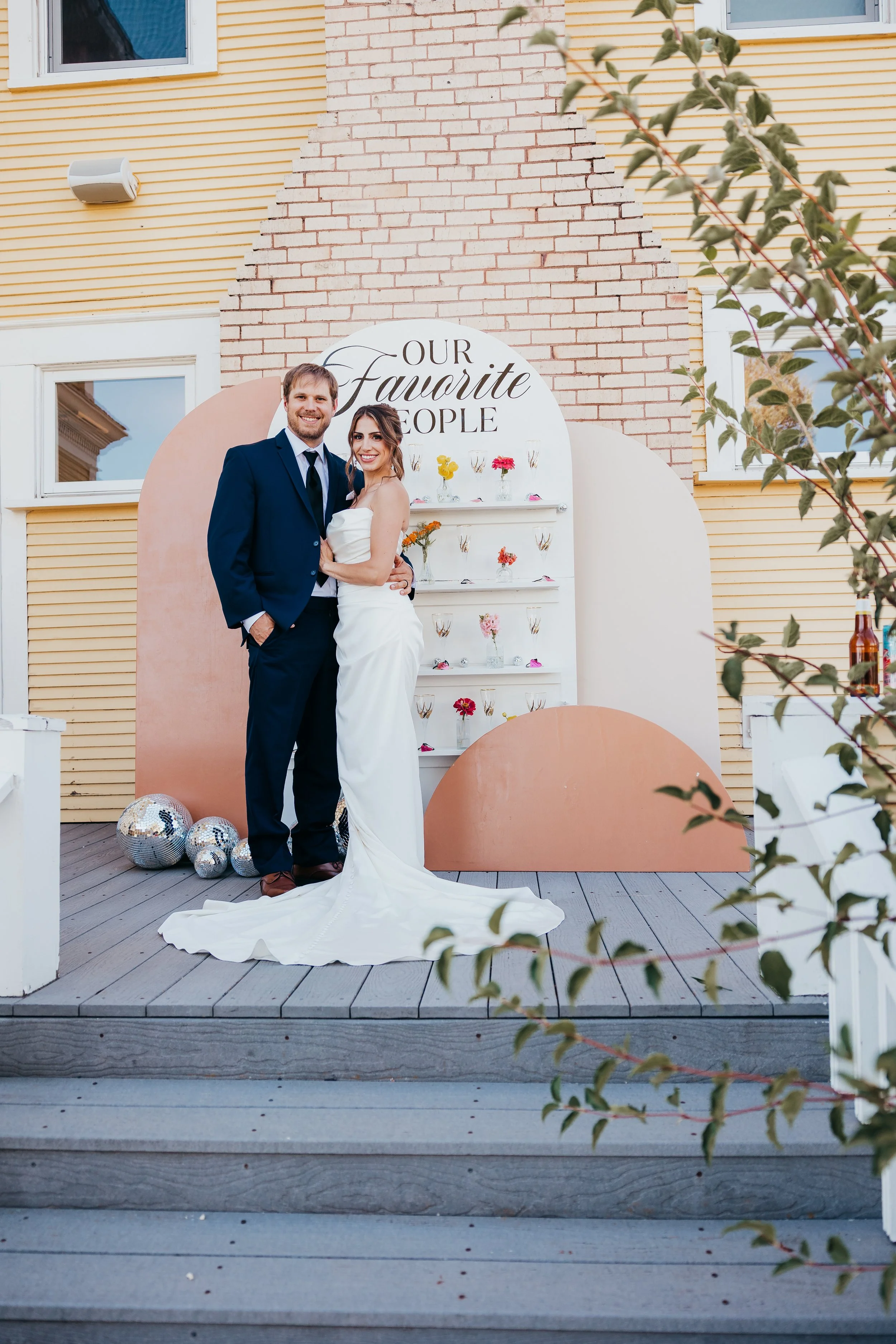 Bride and groom in wedding attire standing on a wooden platform in front of a decorative backdrop that reads 'Our Favorite People,' with shelves holding flowers and small glass containers, surrounded by disco balls, outside a building with yellow sid