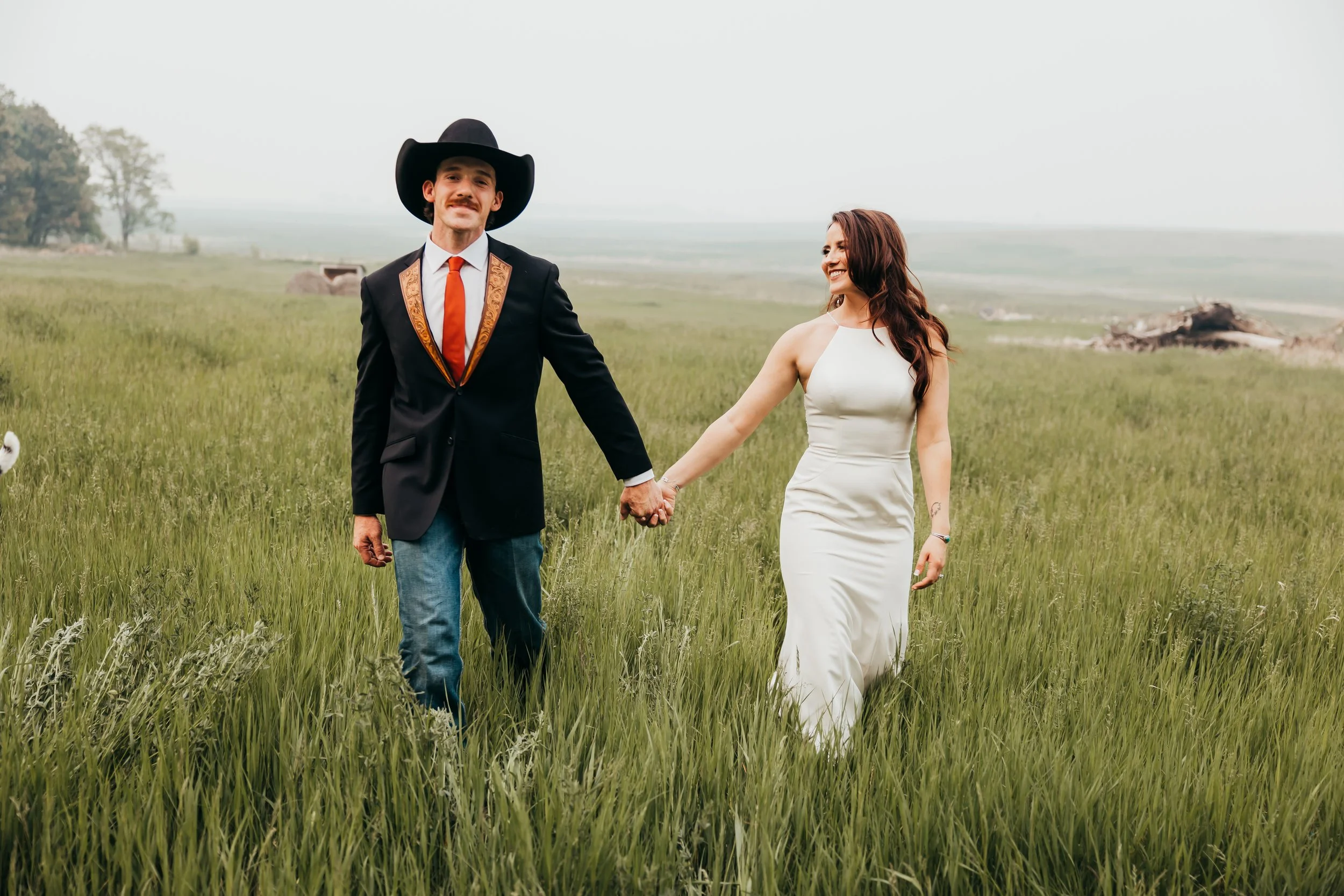 A couple holding hands in a grassy field, the man wearing a dark suit with a large black hat, white shirt, orange tie, and blue jeans, and the woman in a white sleeveless dress, both smiling and looking at each other.