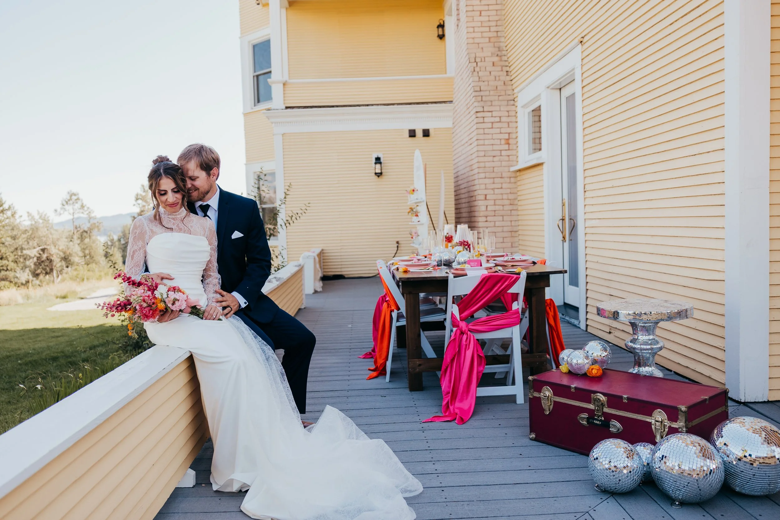 Bride and groom sitting on a ledge on a porch, with a decorated table in the background, celebrating wedding outdoor venue with flowers, disco balls, and decorations.