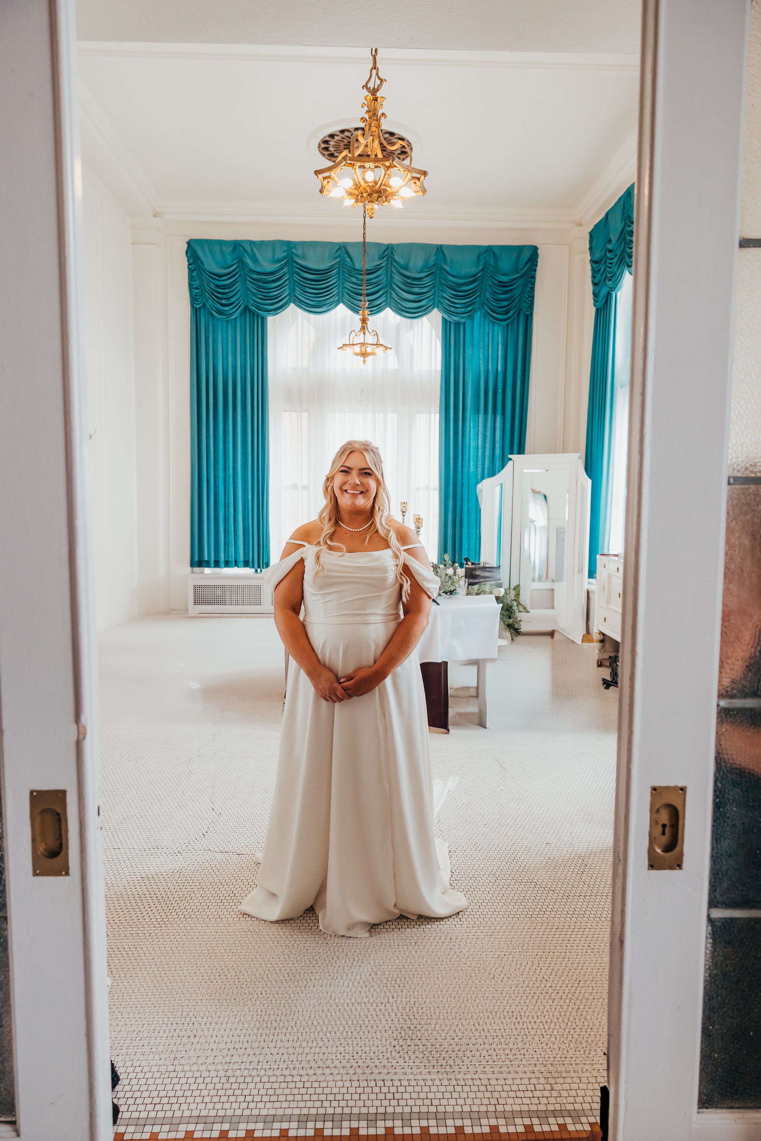 A woman in a white wedding dress smiling in a decorated room with large windows, blue curtains, and vintage decor.