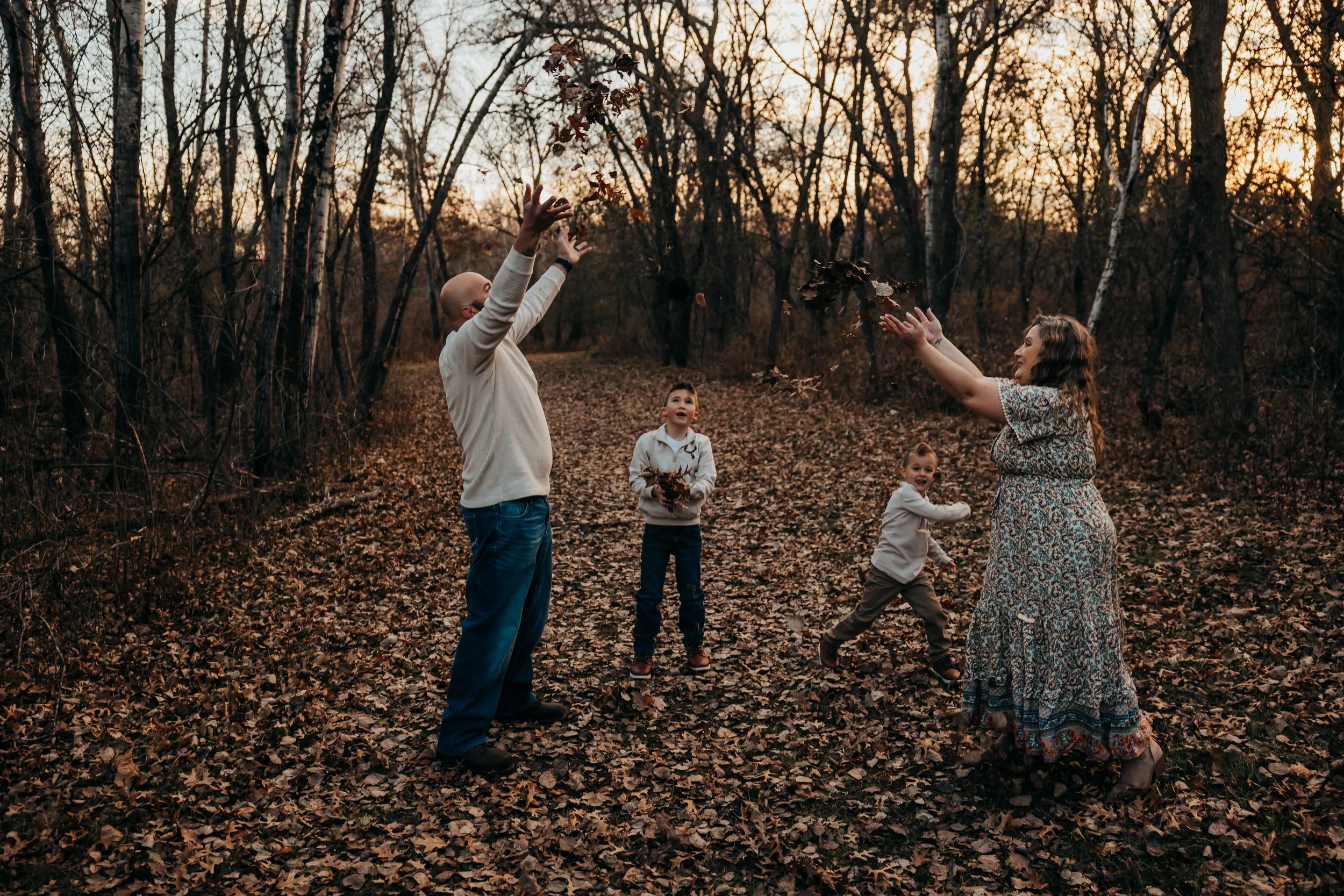 Family of five, including an adult man and woman and three children, playing with fallen leaves in a wooded park during sunset.