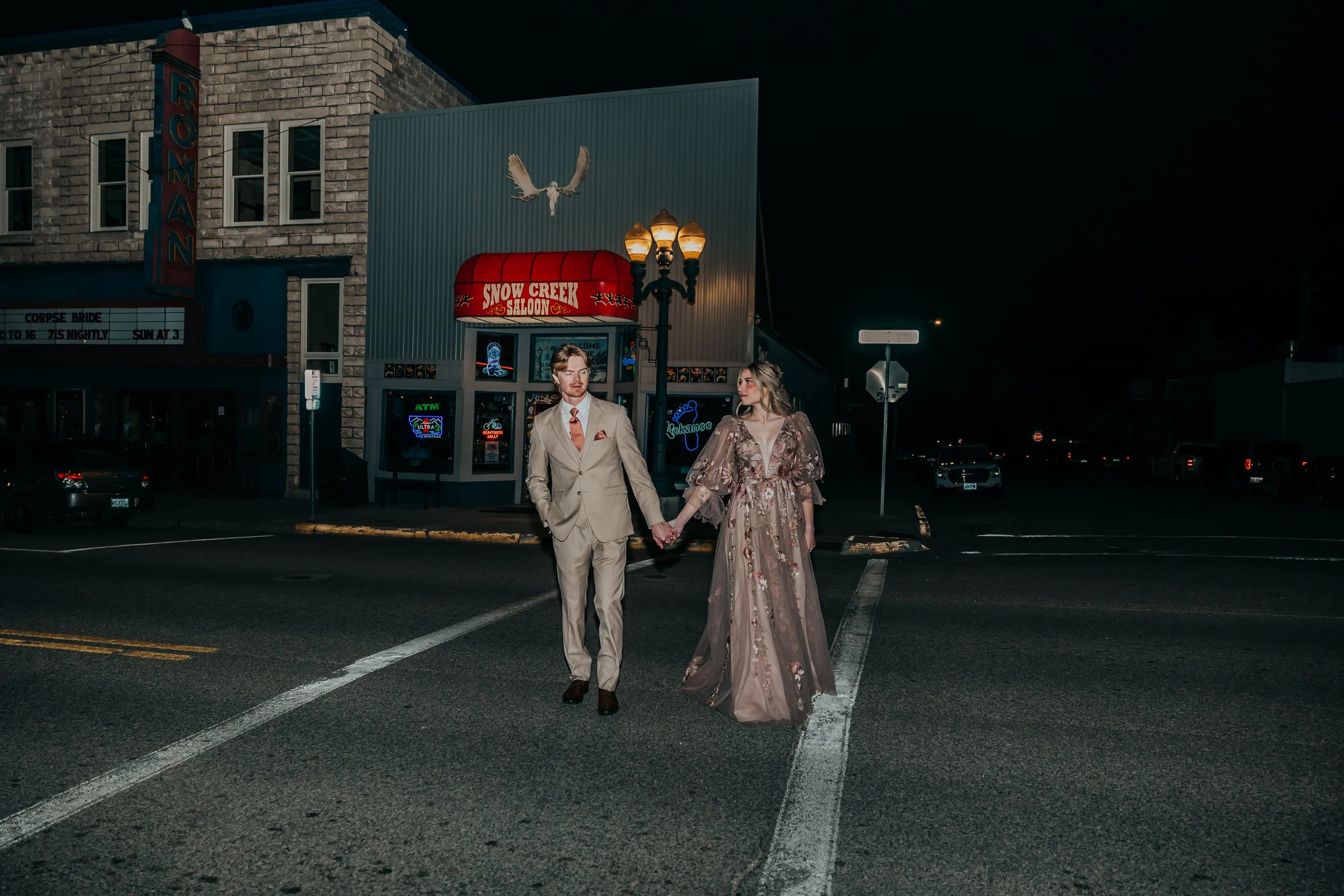 A couple dressed in formal attire holding hands and walking across a city street at night, with a neon-lit storefront and streetlamp in the background.