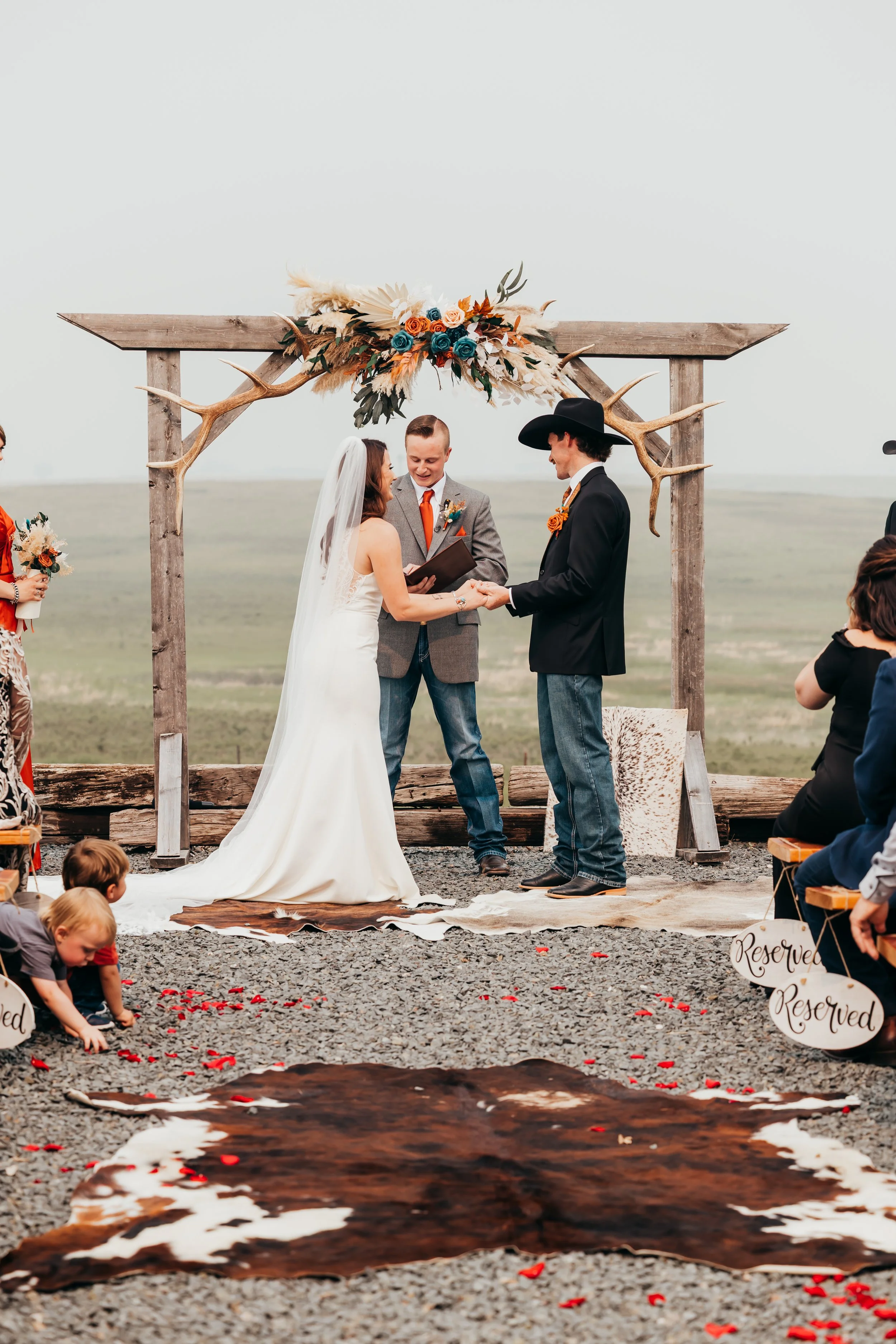 A wedding ceremony taking place outdoors on a gravel surface, with a wooden arch decorated with flowers and antlers. The bride and groom are holding hands and smiling, with the officiant standing between them. Children sit on the sides, and reserved 