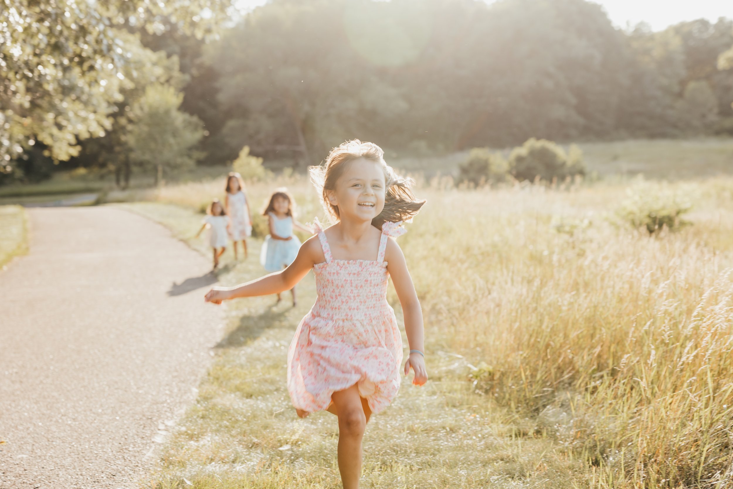 A young girl wearing a pink floral dress smiling and running on a dirt path with several other girls in dresses walking behind her in a sunny outdoor setting with trees and fields.