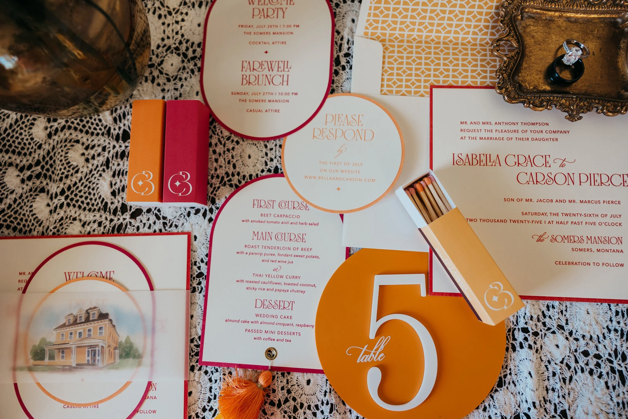 Wedding invitation suite with event details, menu, and table number on lace tablecloth, decorated with matches and jewelry.