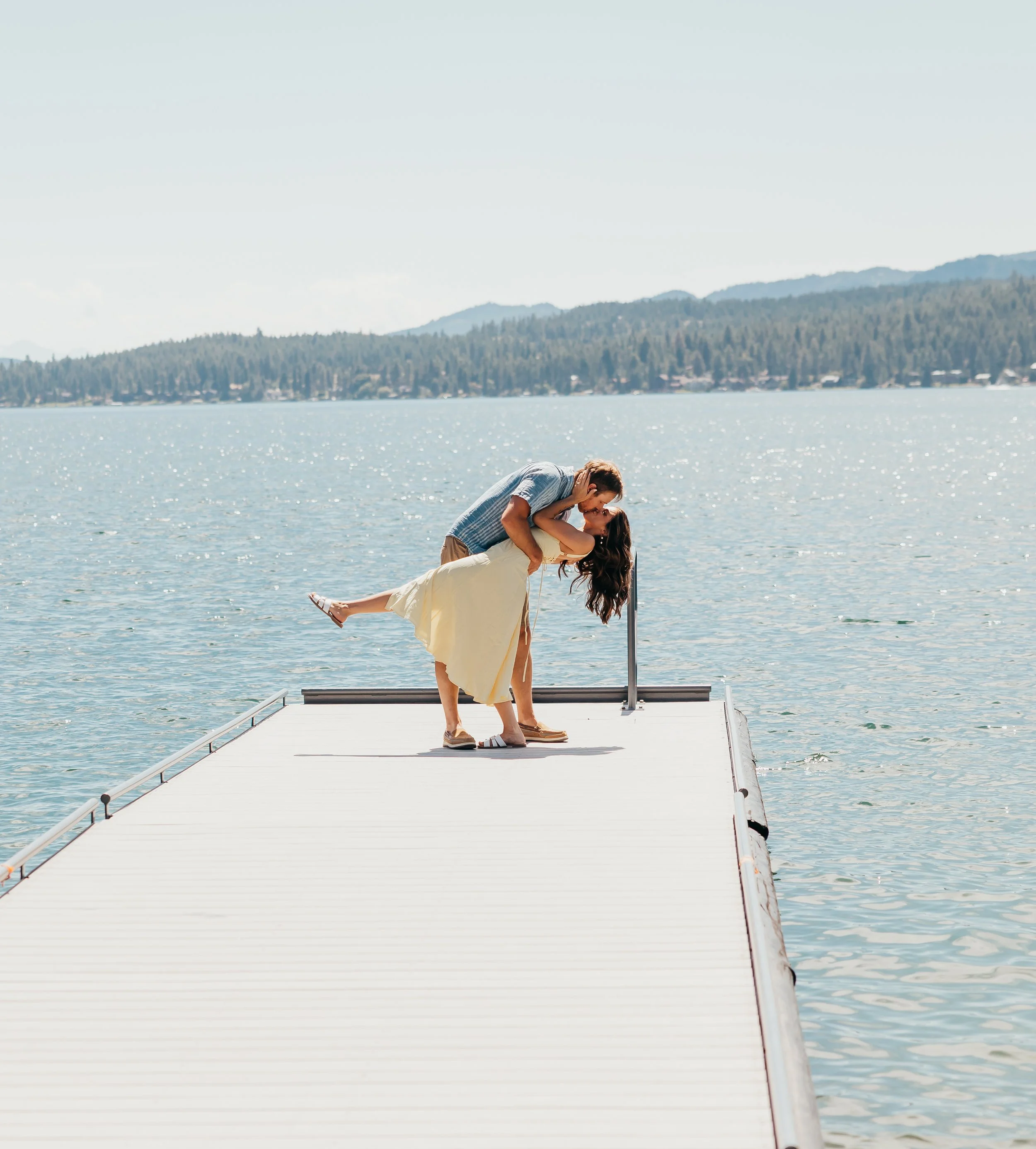 A couple on a dock by a lake, with the man dipping the woman backwards, both smiling and enjoying a sunny day.