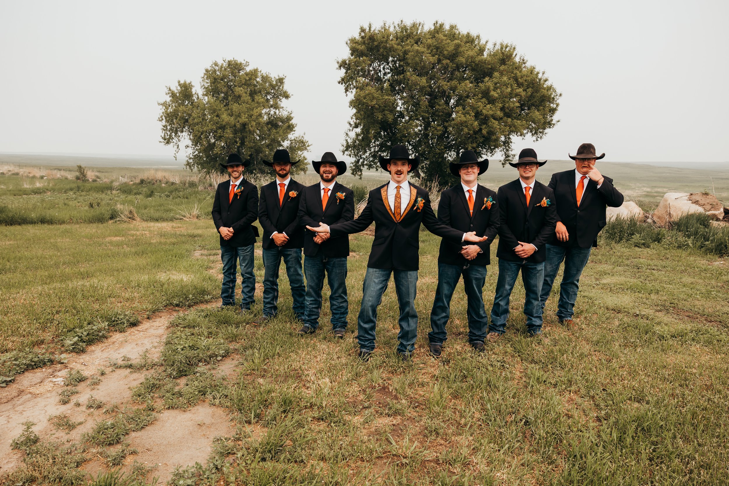 A group of seven men in black suits, orange ties, and black cowboy hats standing on a grassy field with large trees and rocks in the background; one man in front extends his arms open cheerfully.
