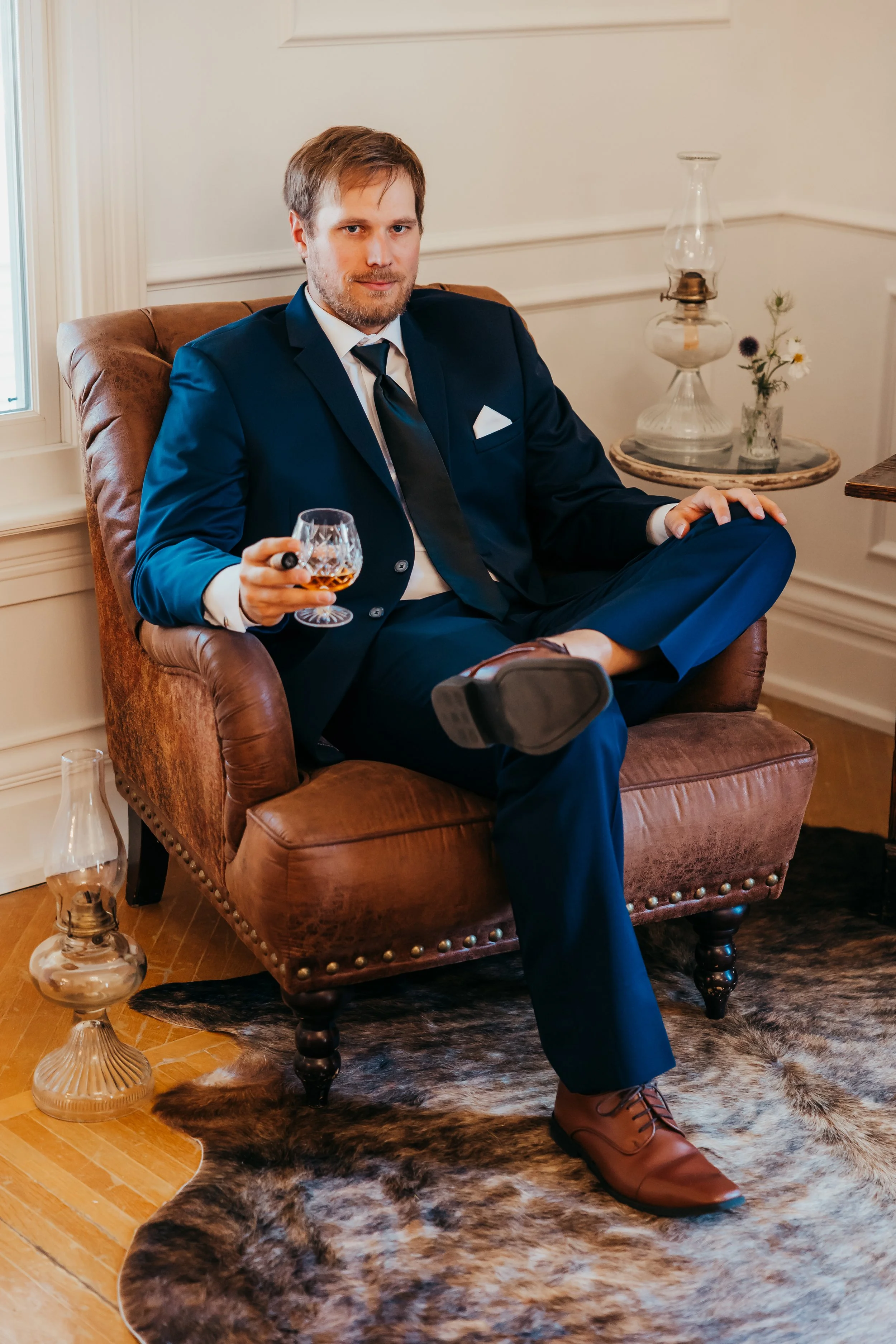 A man in a navy suit sitting on a vintage leather armchair, holding a glass of whiskey, with one leg crossed over the other, in a cozy room with a fur rug and decorative oil lamps.