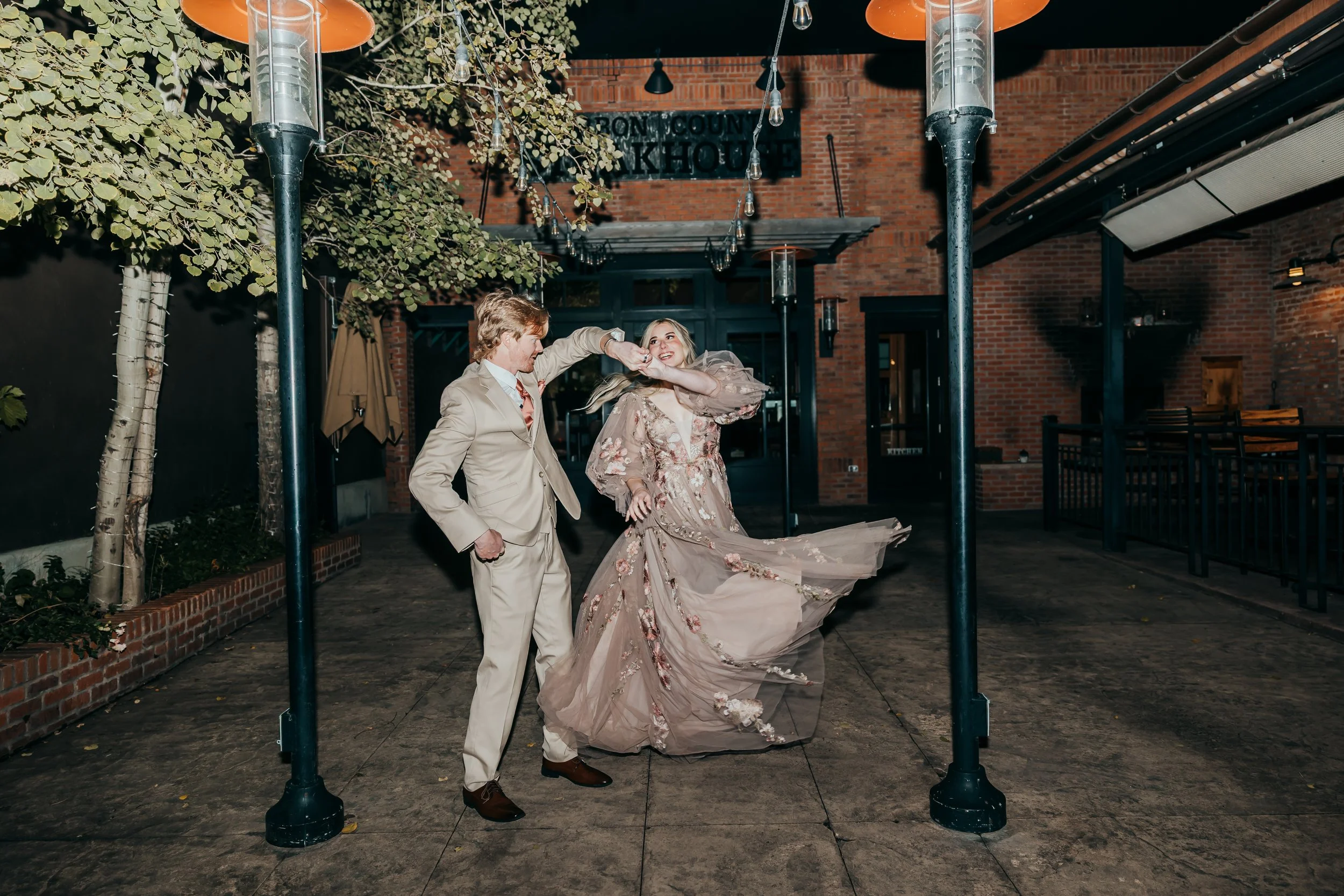 A couple dancing outside at night, with lights and trees around them. The woman is in a flowing, floral dress and the man is in a beige suit.