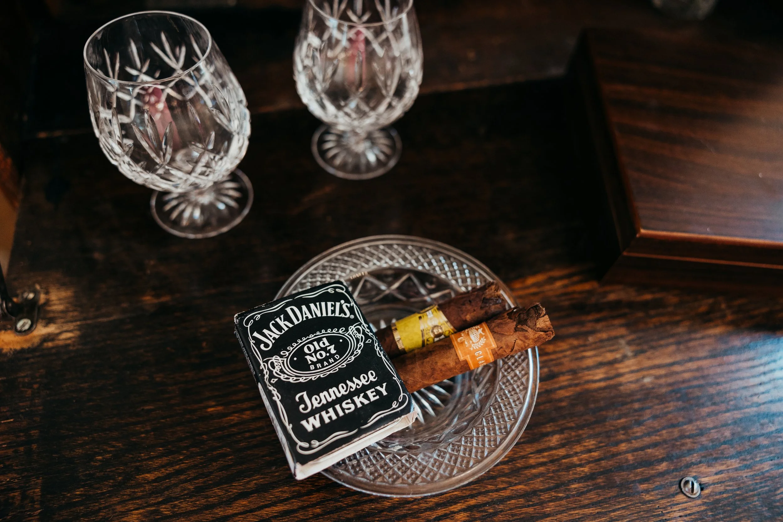 Two empty crystal glasses and a glass ashtray with a pack of Jack Daniel's Tennessee Whiskey and three cigars on a dark wooden surface.
