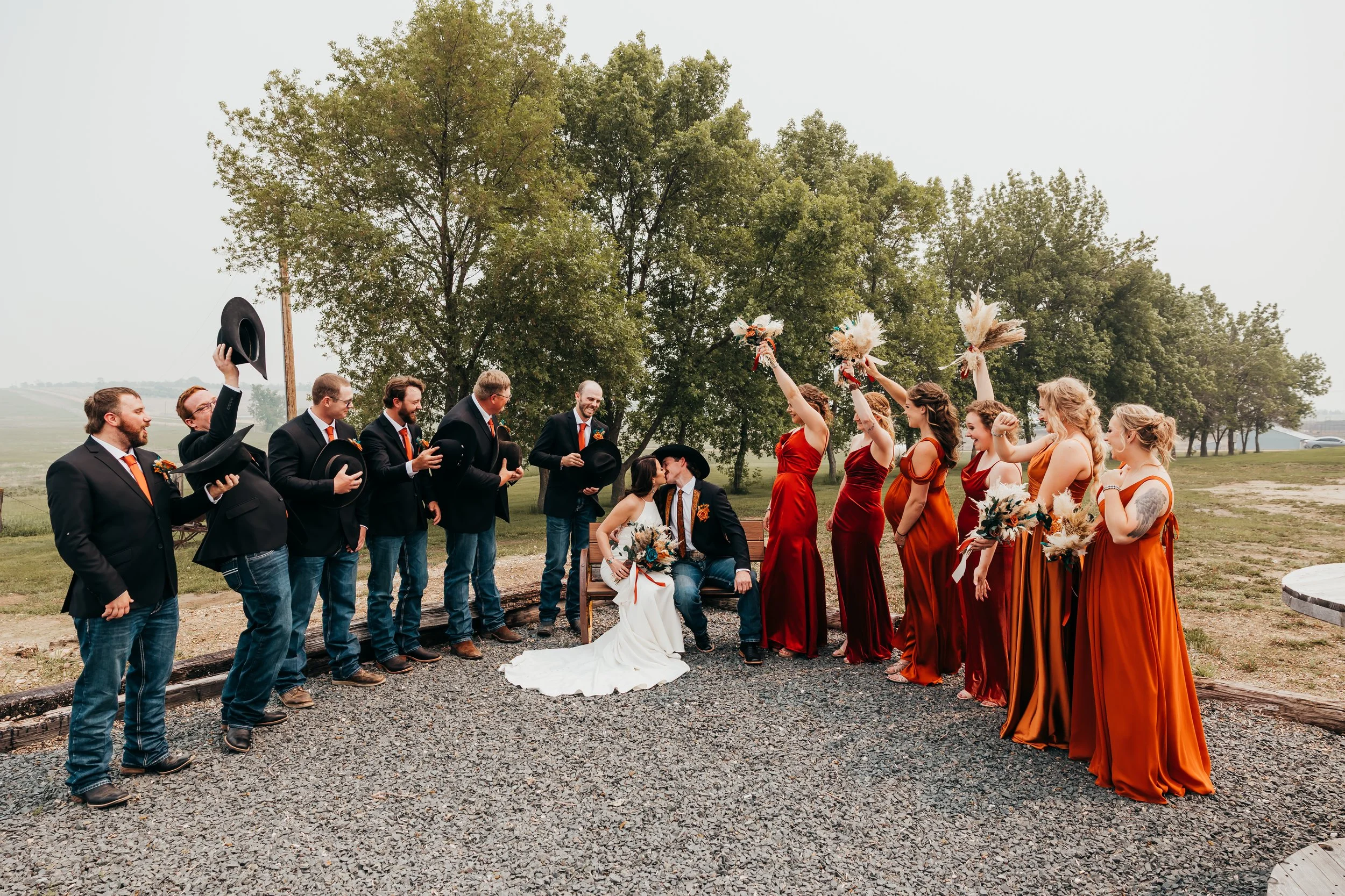 A wedding party with the bride and groom sitting on a bench outdoors, surrounded by bridesmaids in rust-colored dresses and groomsmen in suits with orange ties, celebrating during the day on a cloudy day with trees in the background.