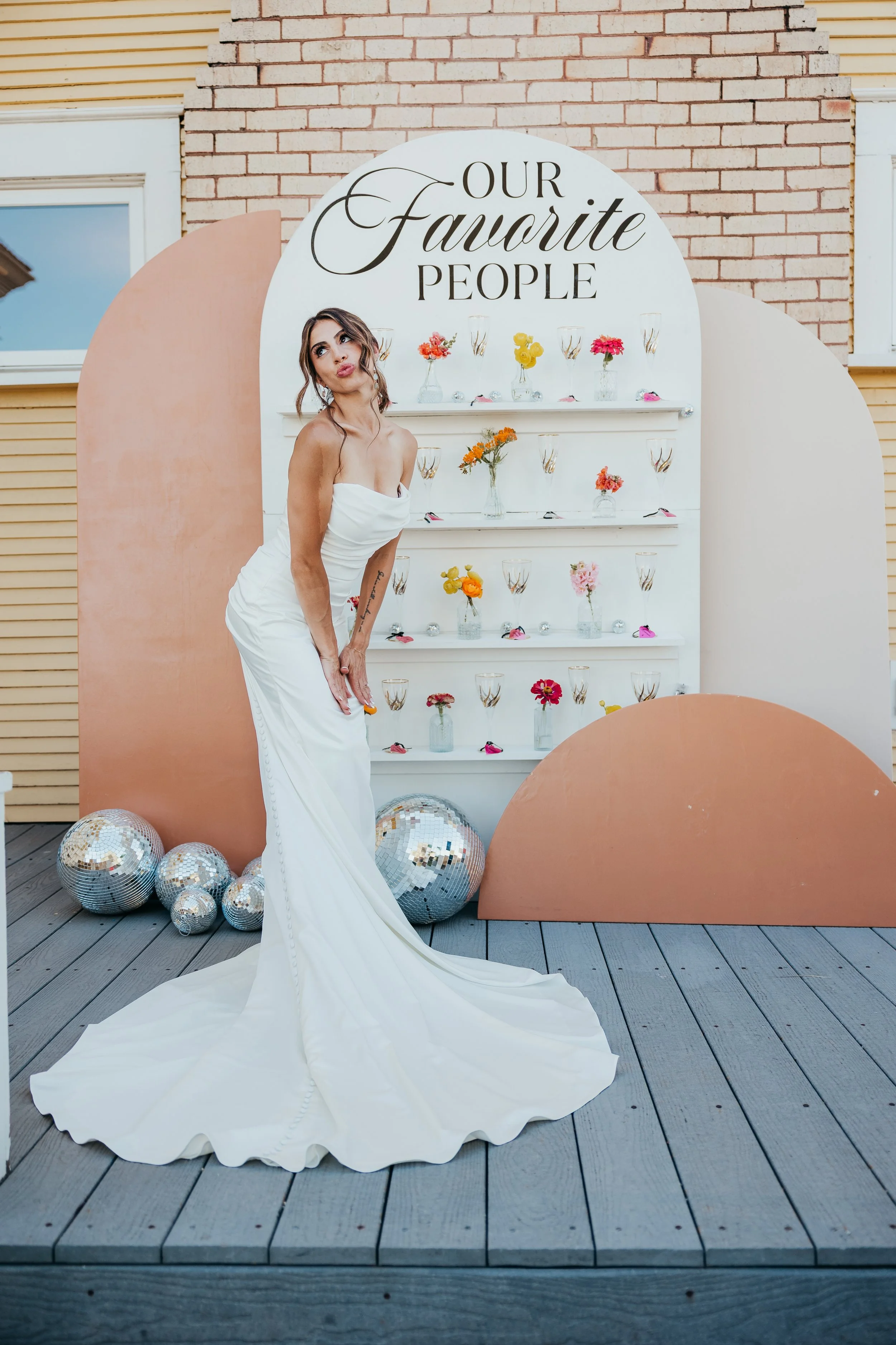 A woman in a white strapless gown poses in front of a display that says 'Our Favorite People,' with colorful flowers and decorative disco balls on the ground.