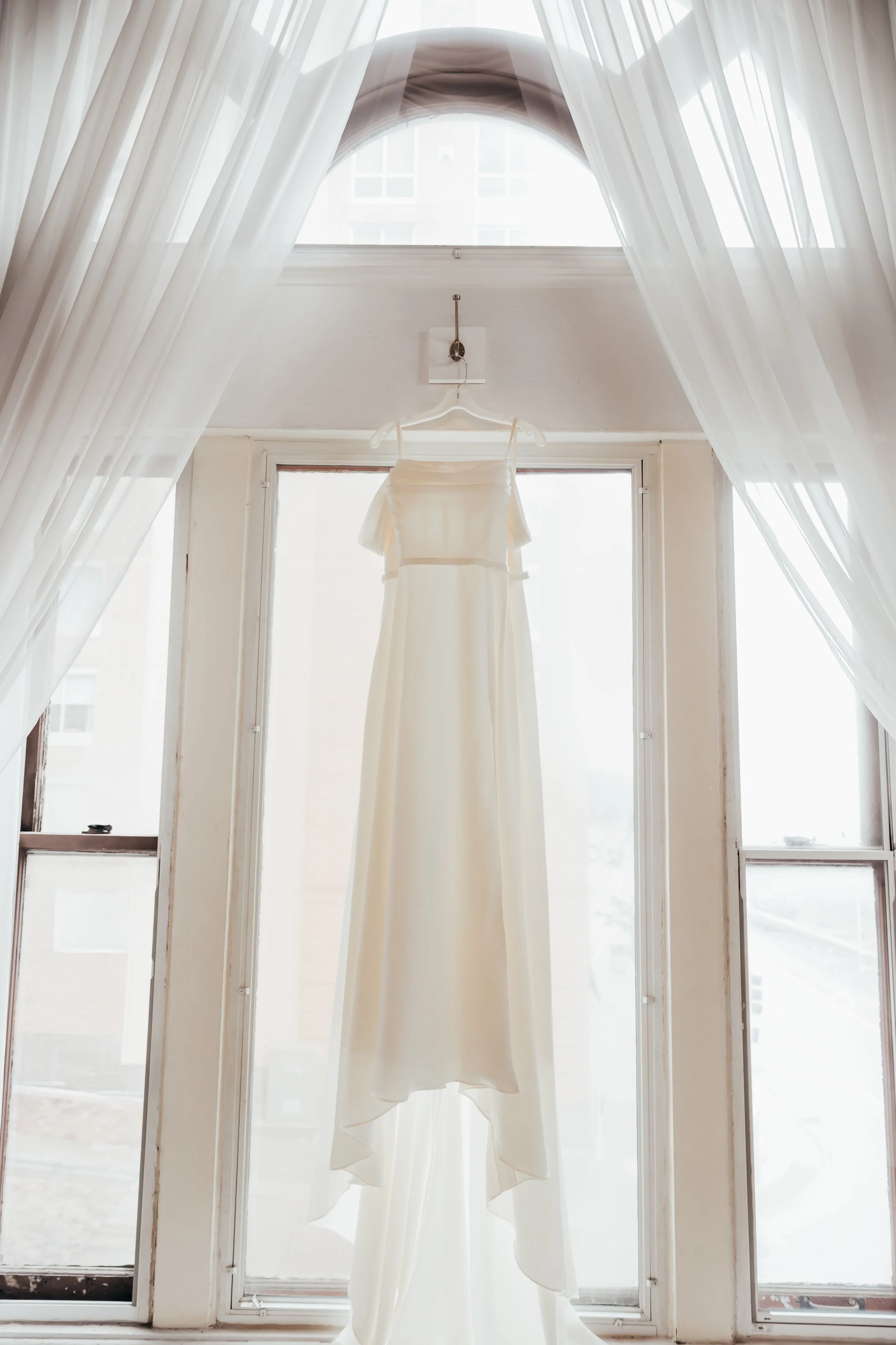 A wedding dress hanging from a hook on a window frame, with sheer curtains on either side, backlit by natural light.