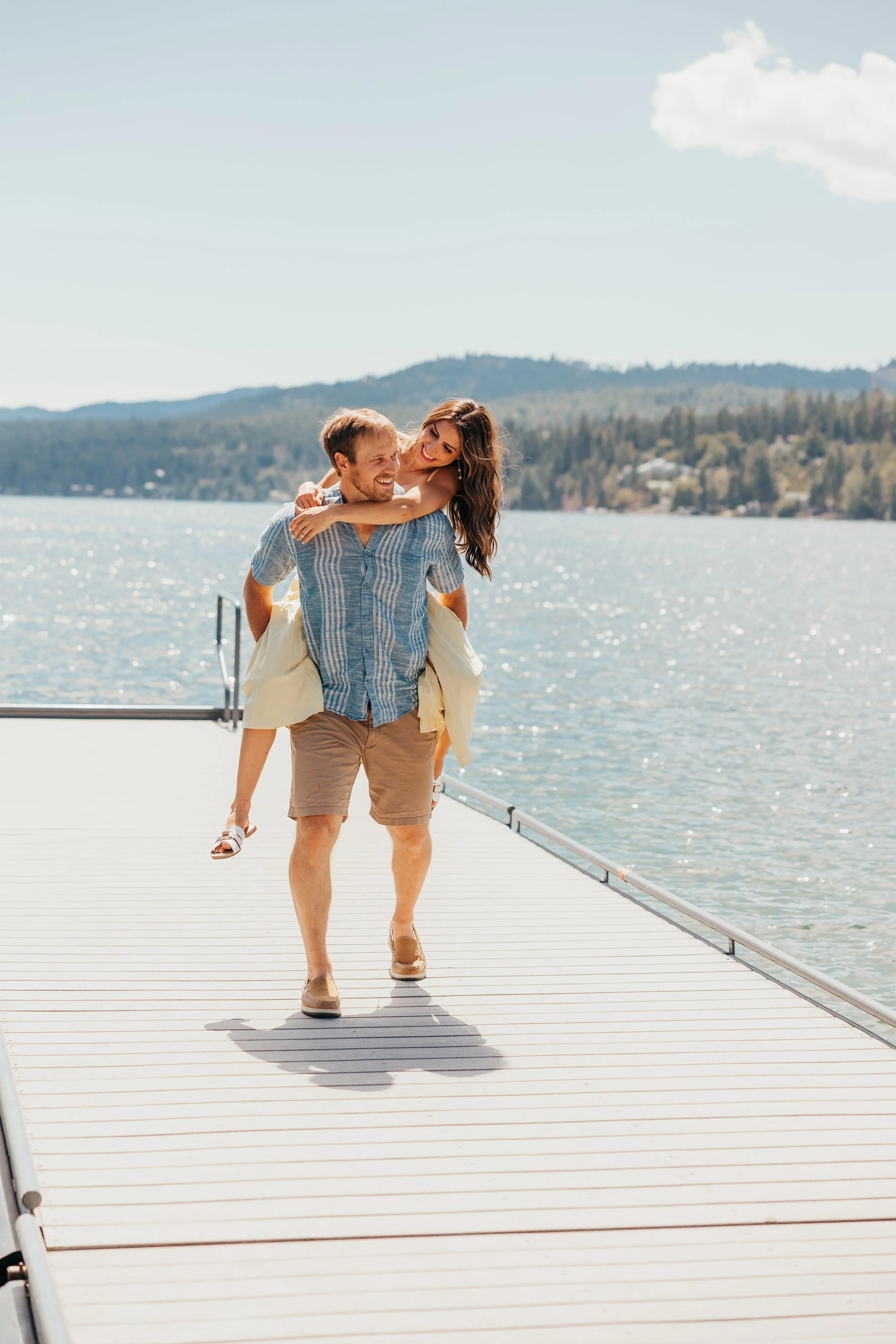 A couple is smiling and enjoying time together on a dock by a lake on a sunny day. The woman is giving the man a piggyback ride, and they are dressed in casual summer clothes with mountains in the background.