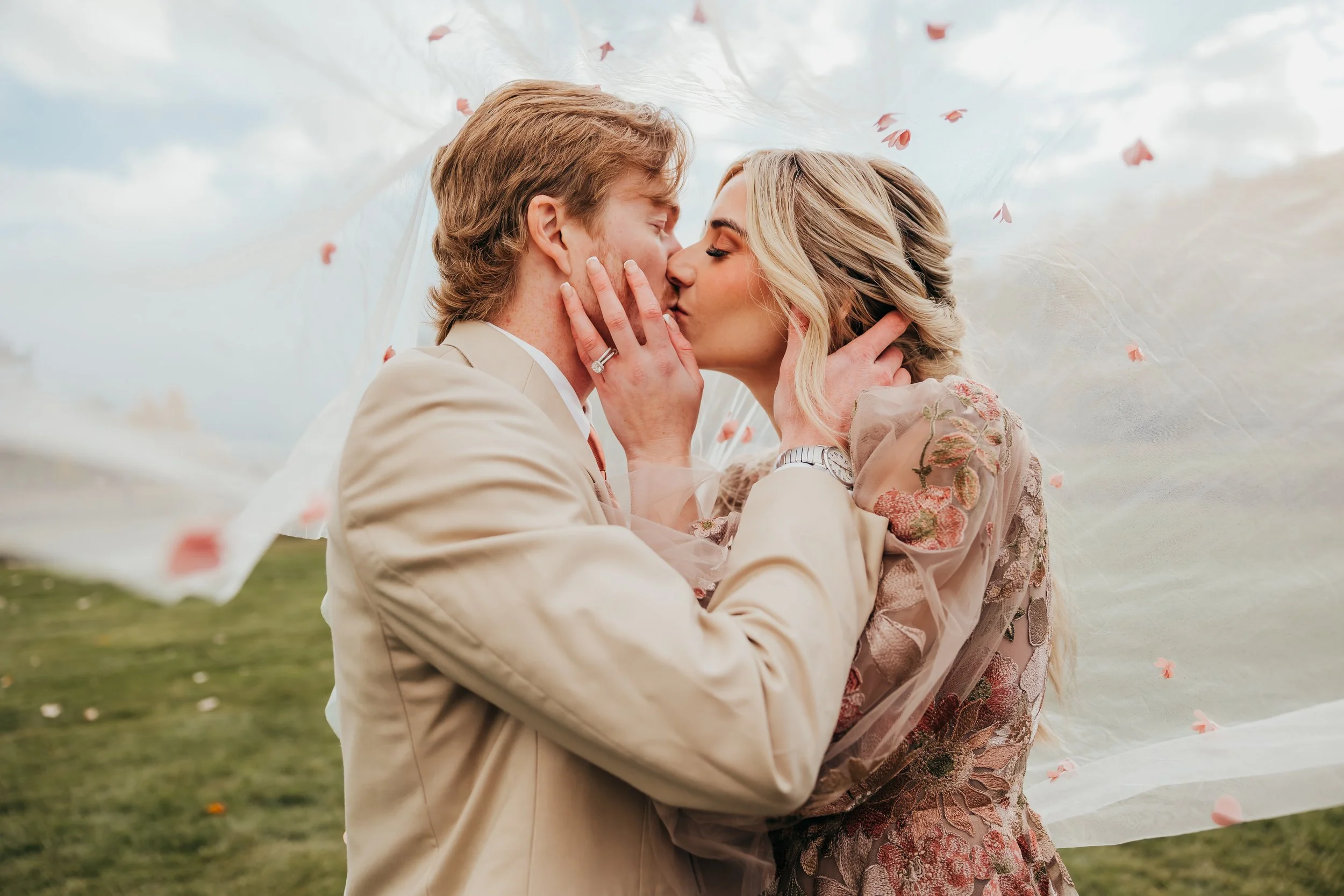 A romantic couple sharing a kiss outdoors with a veil blowing in the wind, surrounded by a grassy landscape and cloudy sky.