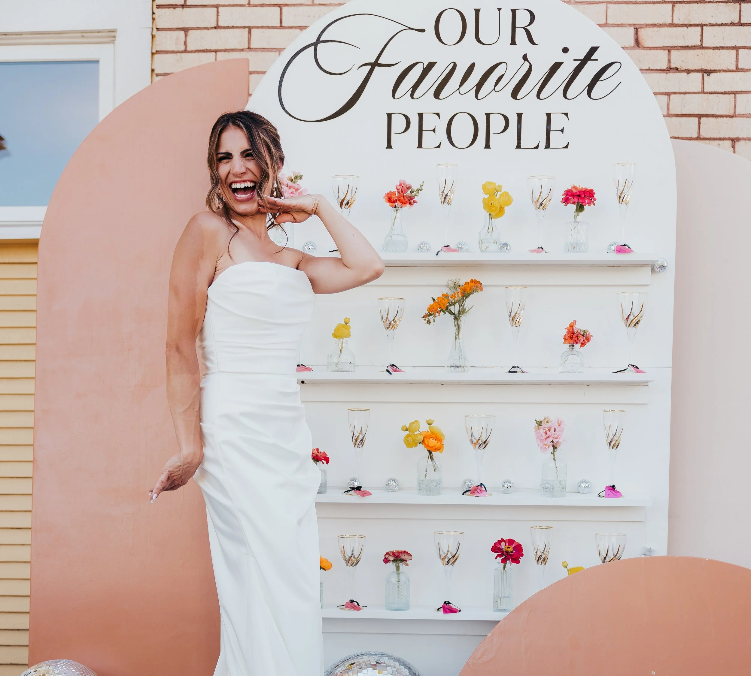 A woman in a white strapless wedding dress smiling and posing in front of a decorative display with flowers in glass vases and champagne glasses. The display has a sign that reads "Our Favorite People."