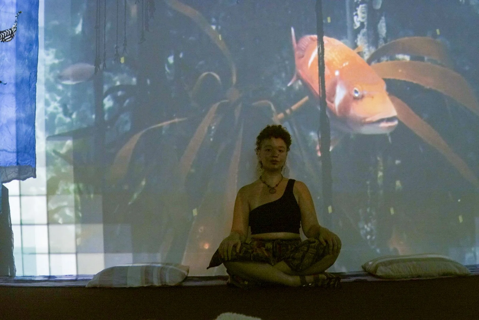 Person sitting cross-legged on a platform with cushions, in front of a projection of an underwater scene with large orange fish and background aquatic scenery.