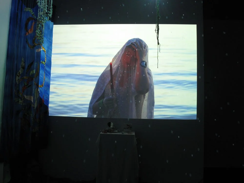 Person dressed in white with a veil, standing by water, holding a candle, visible on a screen in a dark room.