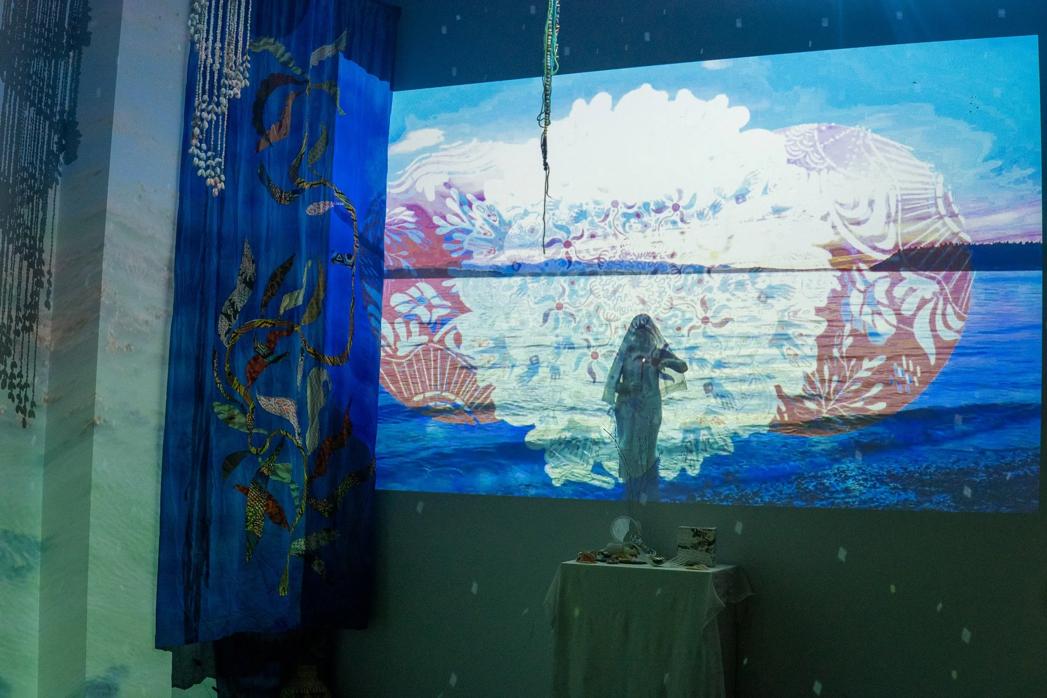 An art installation featuring a large projection of a landscape with clouds and water, a traditional patterned curtain on the left, and a small table with decorative items on the right.